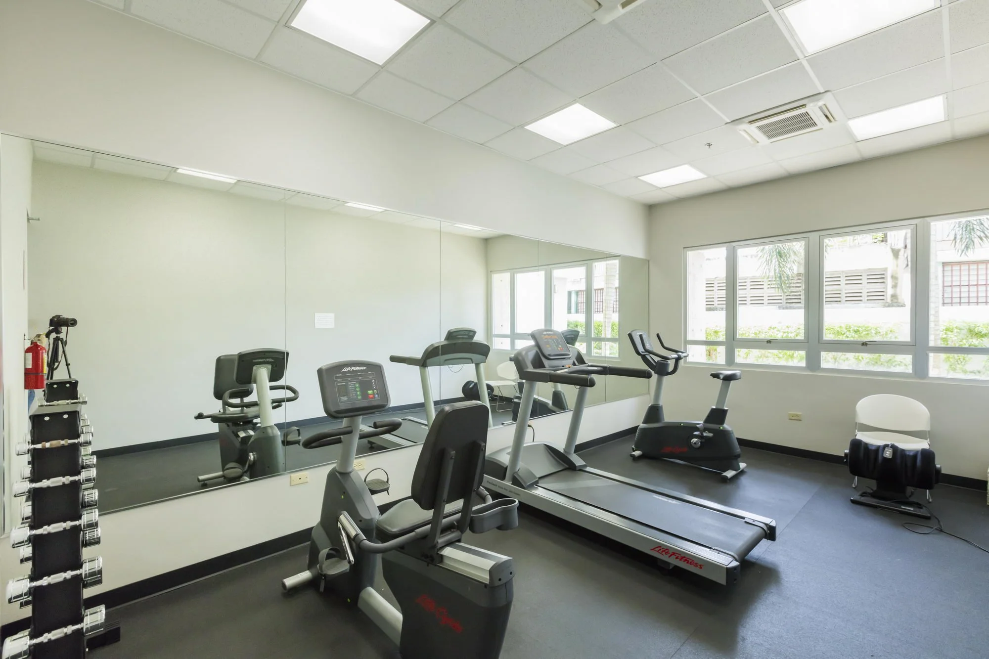 Fitness center with treadmills, elliptical machines, and a mirror along one wall, with large windows providing natural light.