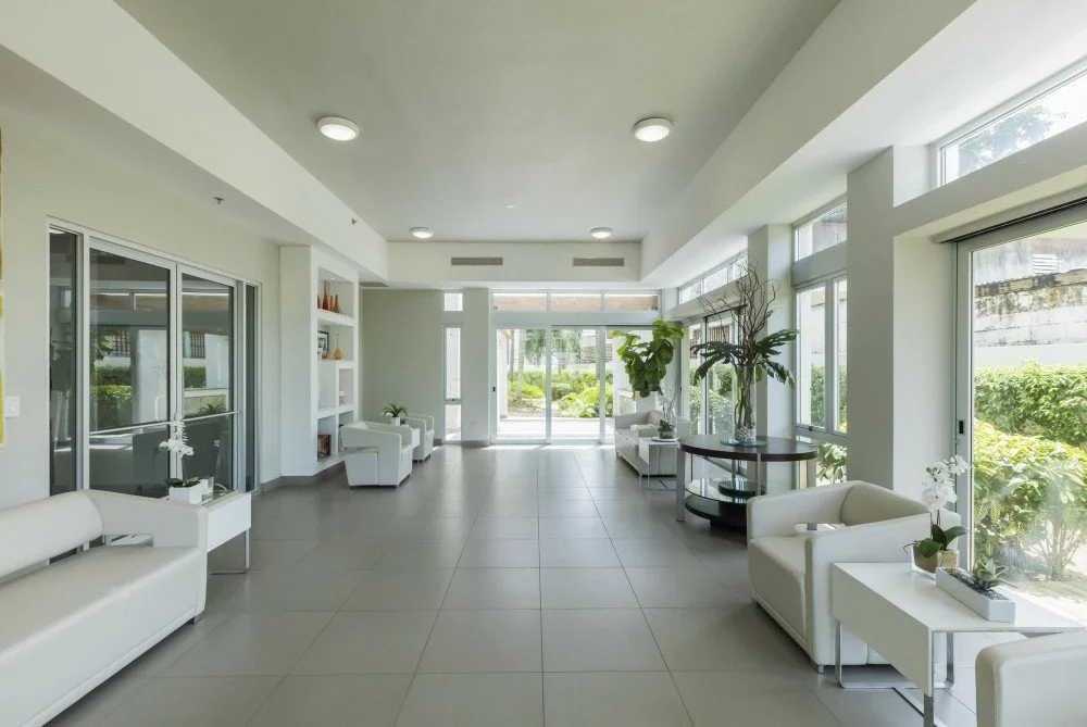 Spacious modern lounge area with white sofas, large windows, potted plants, and minimalistic decor.