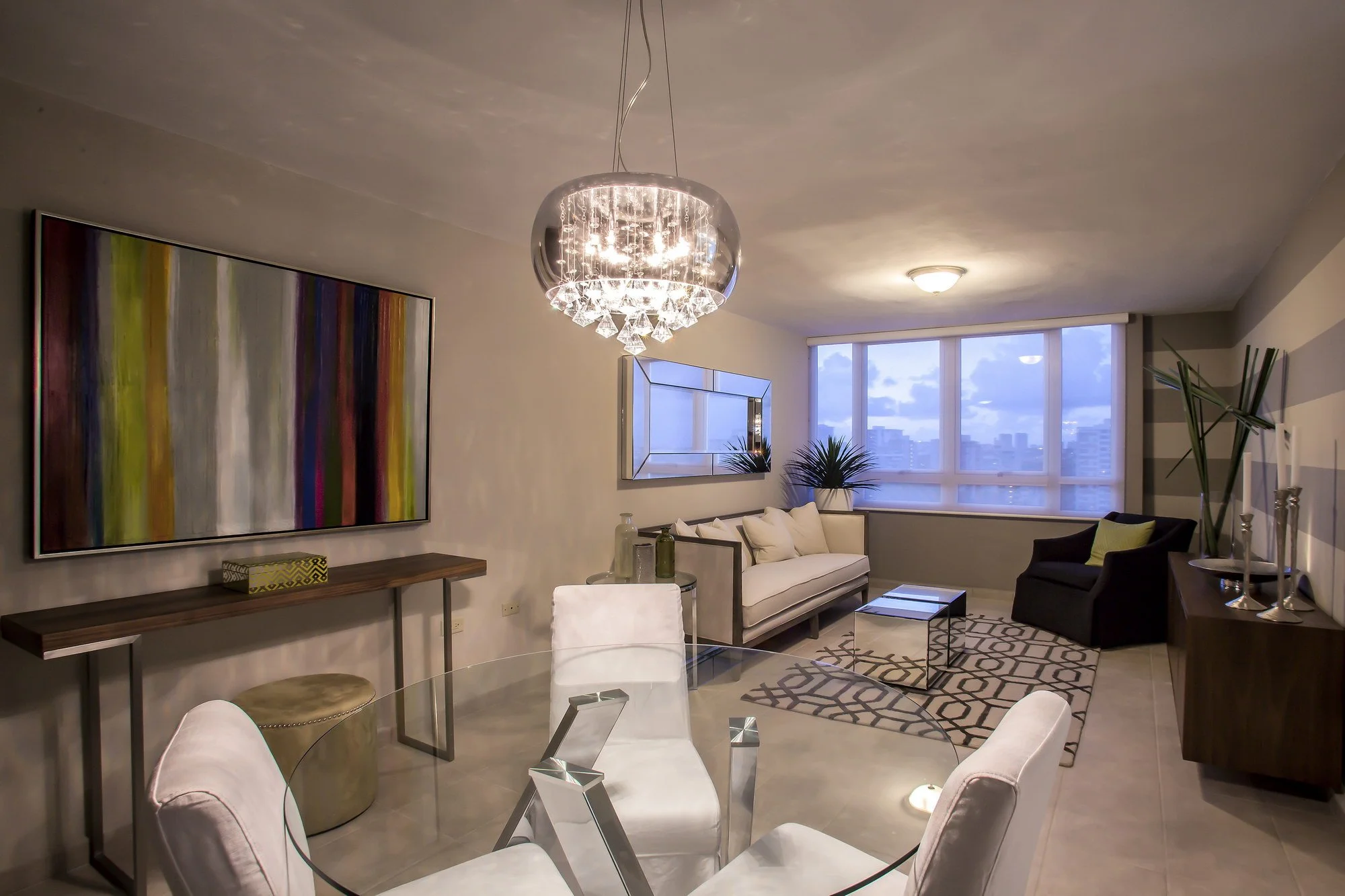Living room with modern furniture, a large abstract painting on the wall, a chandelier, and large windows showing city buildings outside.