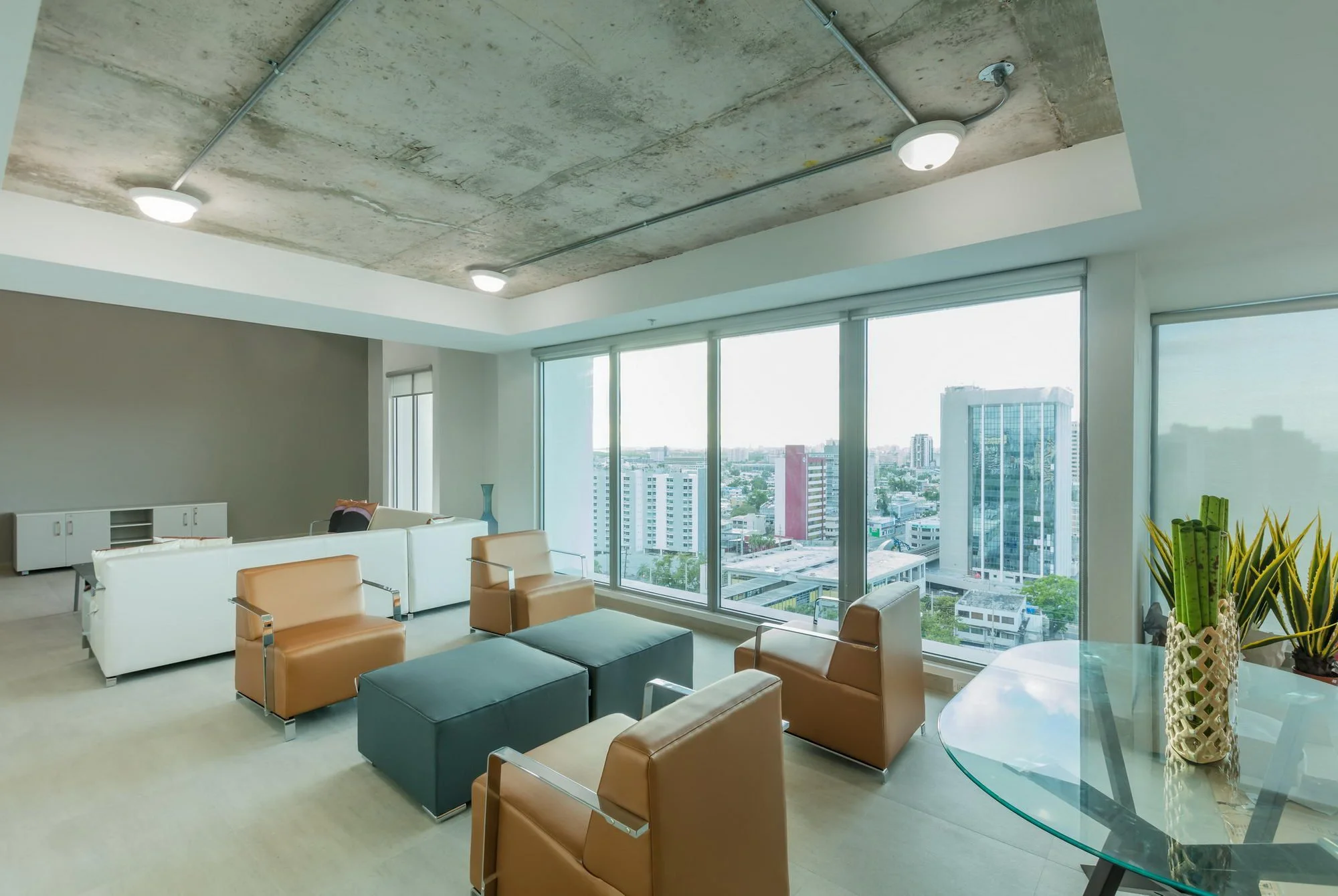 Modern office lounge with large windows, city view, leather chairs, white sofas, potted plants, concrete ceiling, and ceiling lights.