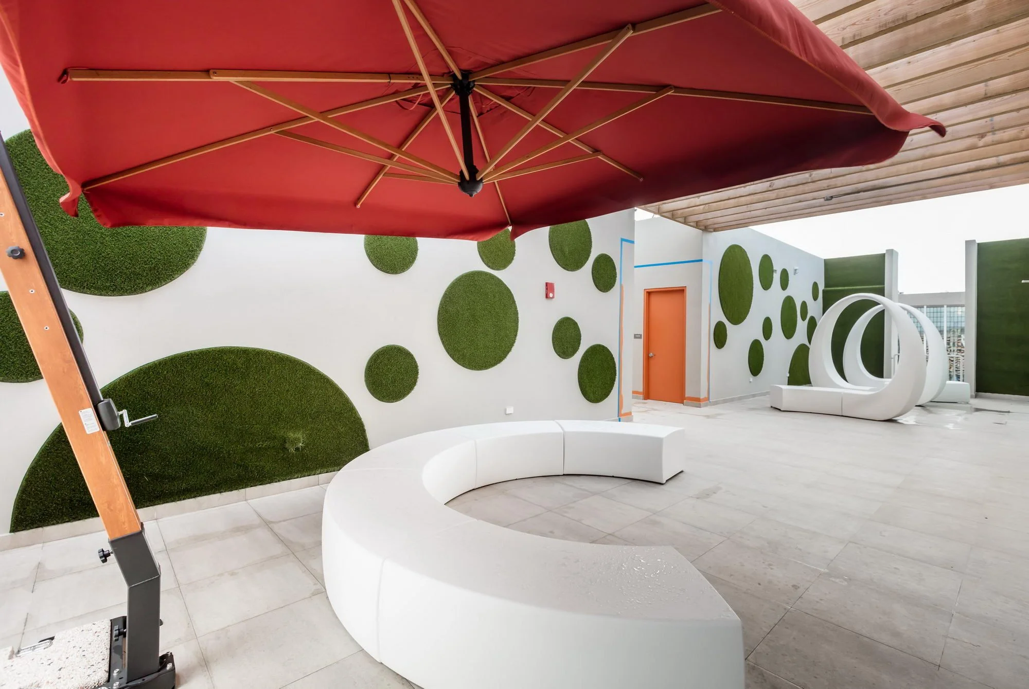 Outdoor modern seating area with white circular benches and green circular wall decorations, a large red patio umbrella, and a white chimney-shaped sculpture or fixture.