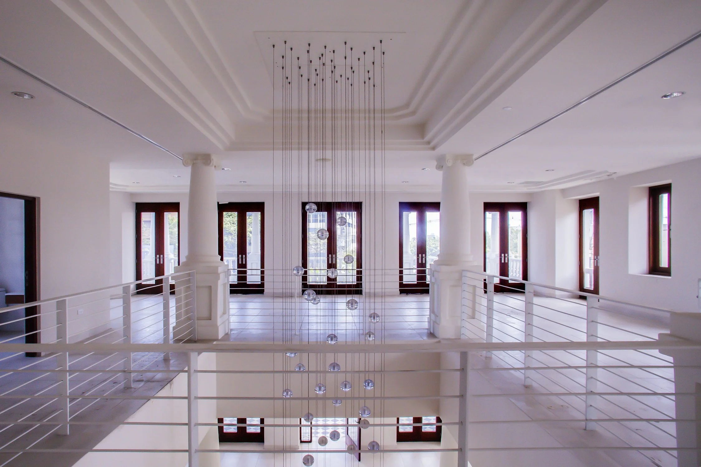 A spacious modern interior with white walls, large windows, white columns, and a high ceiling featuring a hanging light fixture. The area is illuminated by natural light from multiple windowed doors, with a staircase railing visible in the foreground
