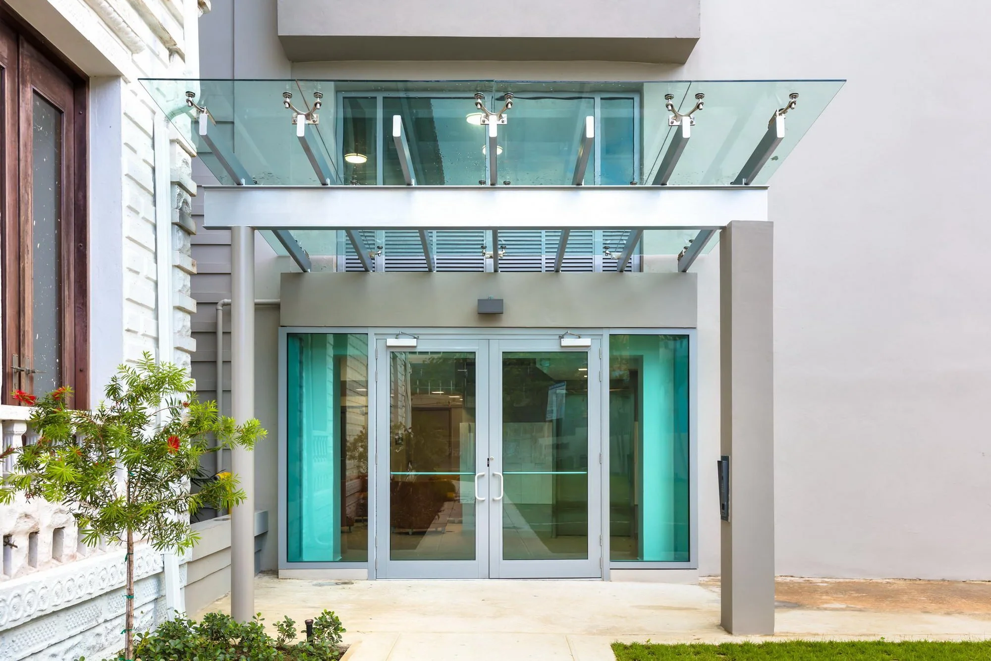 Modern building with glass balcony and sliding glass doors, some greenery and flowering plants in front.