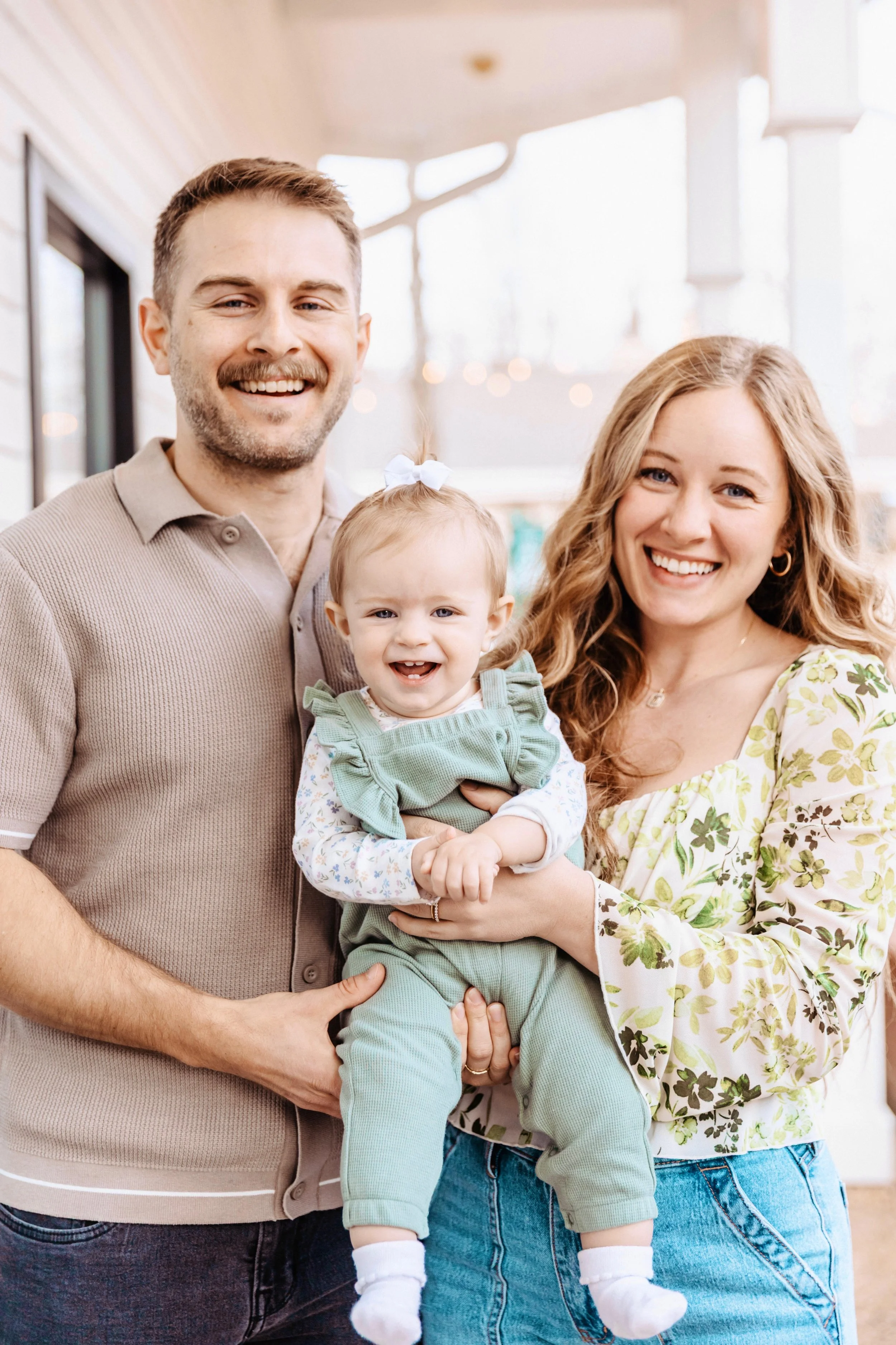 Smiling family of three, with a man, woman, and a toddler girl, standing indoors near large windows with sunlight, all dressed casually and happy.