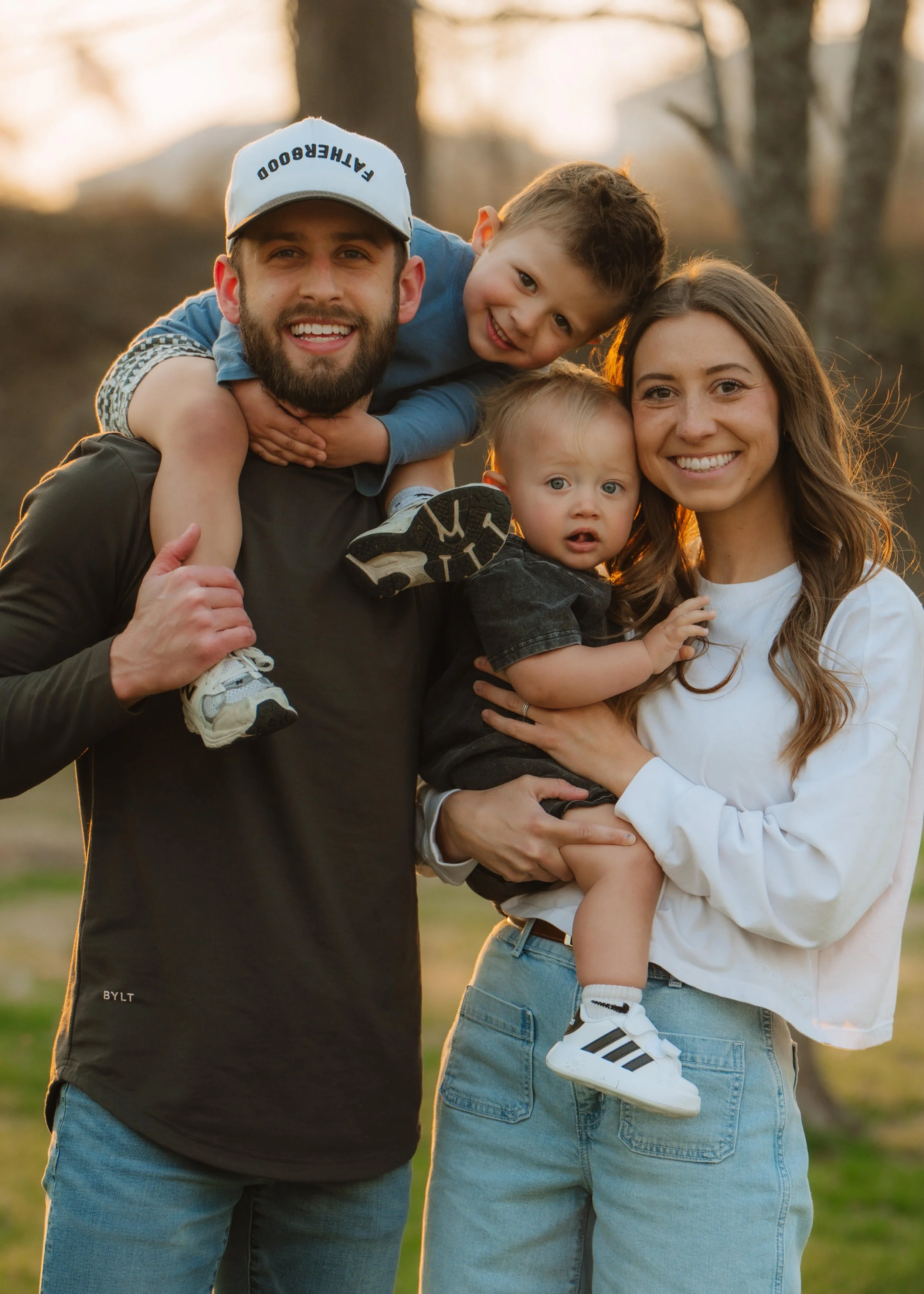 A happy family outdoors during sunset, with a man carrying a young boy on his shoulders, and a woman holding a toddler, all smiling at the camera.