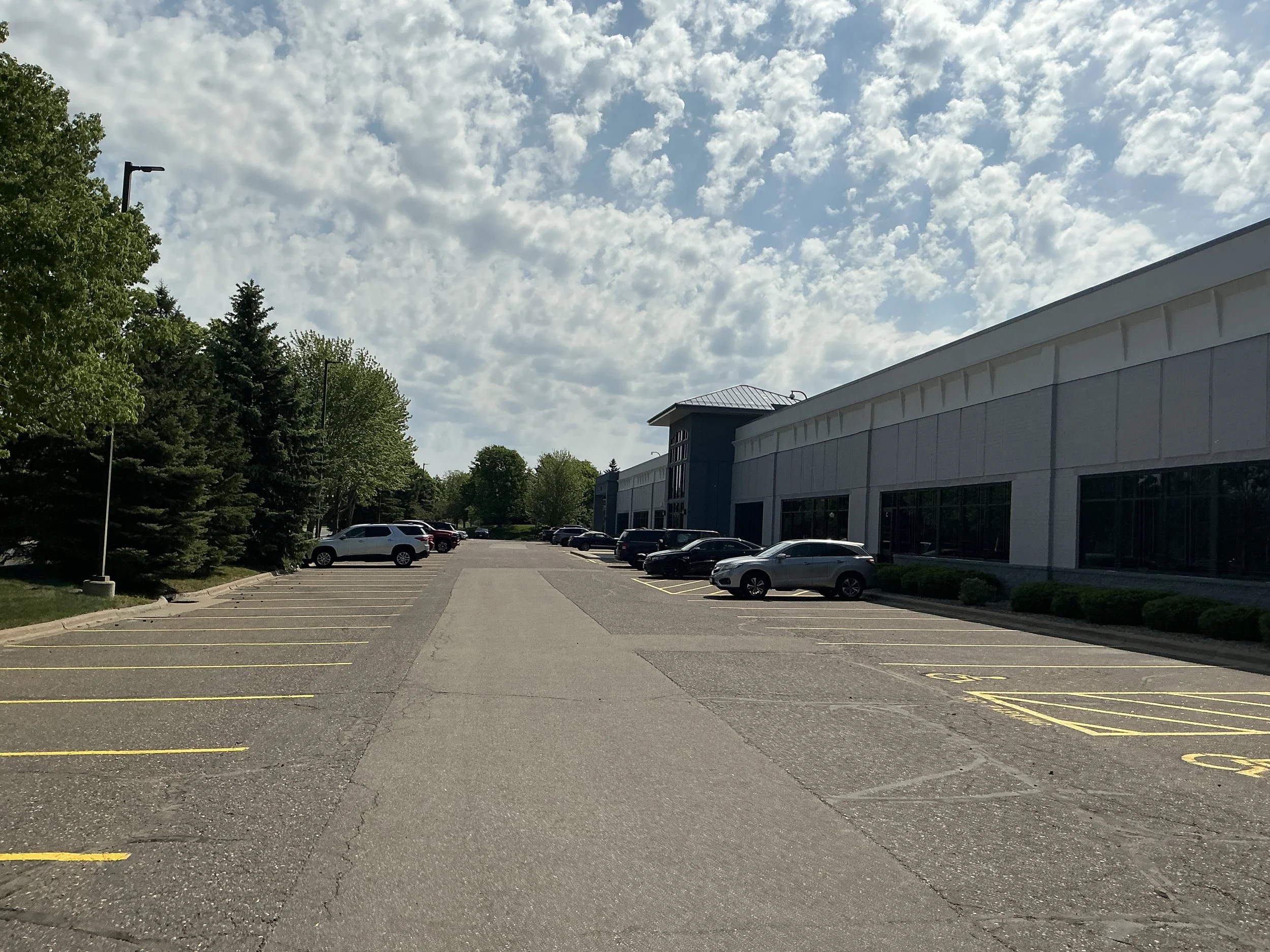 Empty parking lot with marked parking spaces and a large white building on the right, green trees on the left, and a partly cloudy sky overhead.