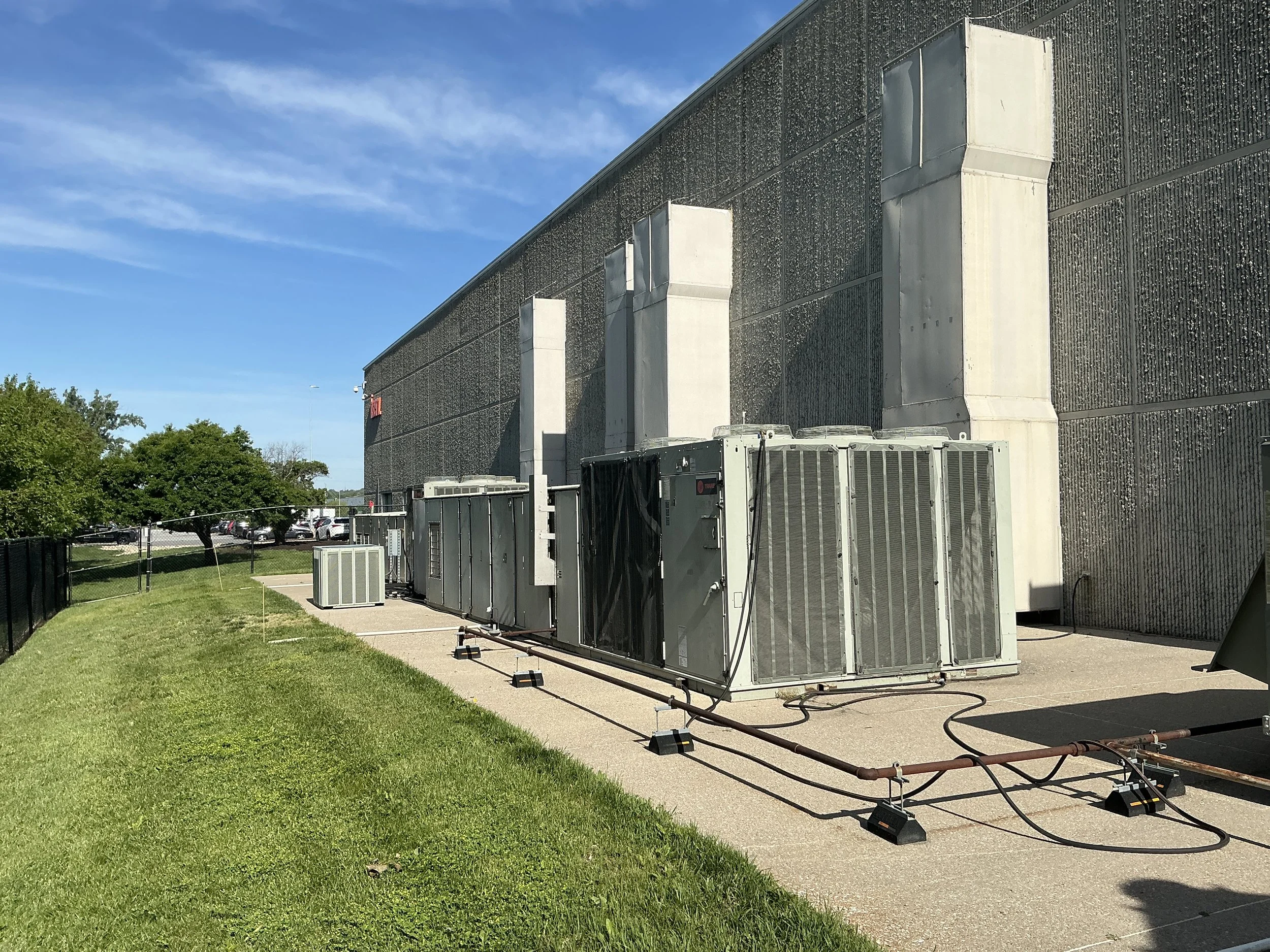 Exterior view of a large commercial building with dryer vents on the exterior wall, air conditioning units and pipes on the ground, and trees and parking lot in the background under a blue sky.