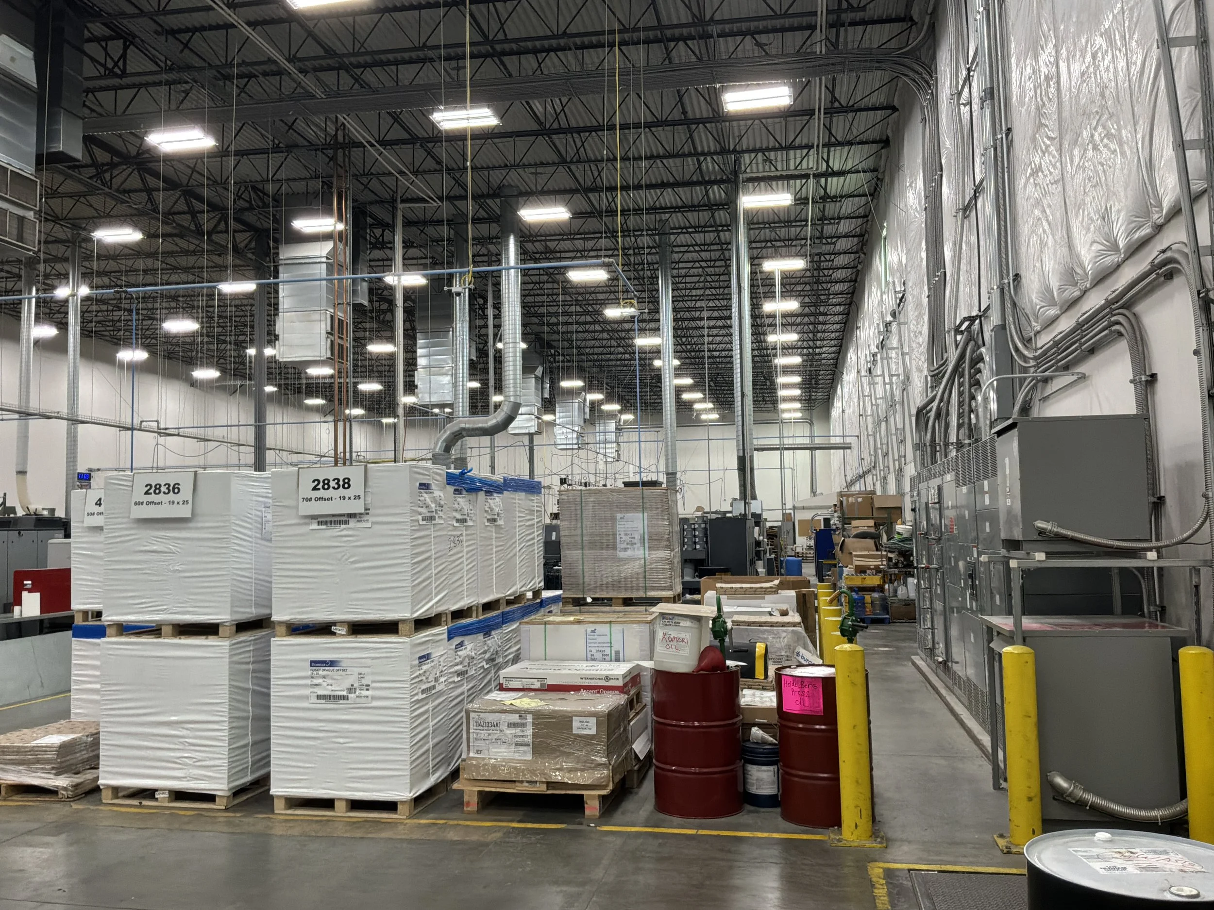 Inside a large warehouse with stacks of large white packages on pallets, exposed ductwork and electrical conduit along the walls, and bright ceiling lights.