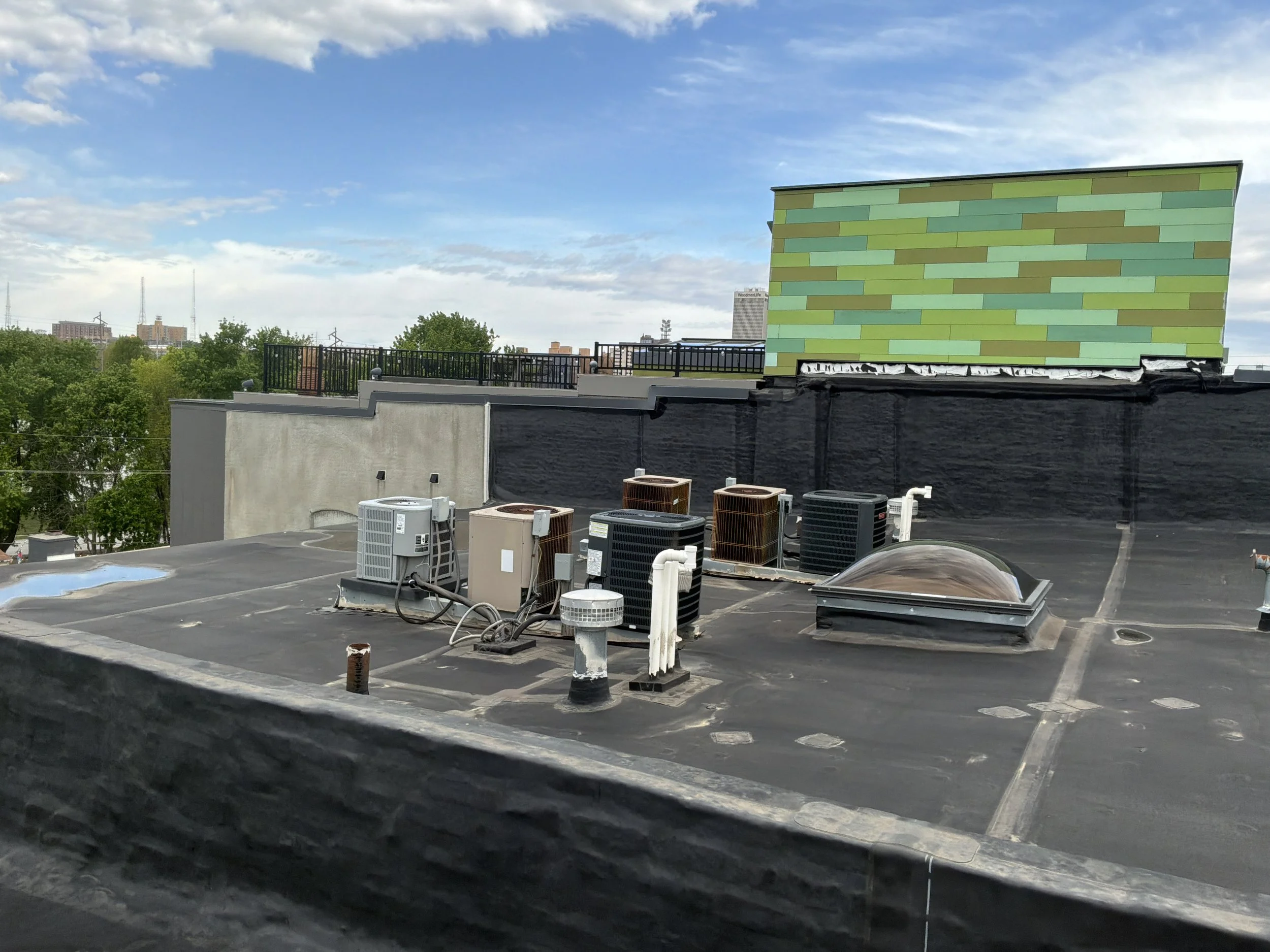 Rooftop view showing HVAC units, a black roof with some debris, and a decorative wall with green and yellow brick pattern. In the background, trees and buildings under a partly cloudy sky.