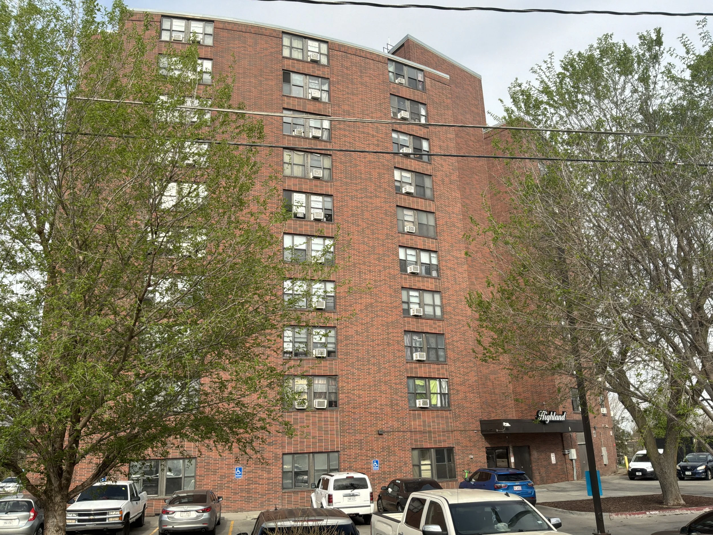 A tall red brick apartment building with multiple windows and air conditioning units, partially obscured by trees with green leaves. The building has a parking lot in front with several parked cars and handicapped parking signs, and a small sign with the word 'Highland' at the bottom right.