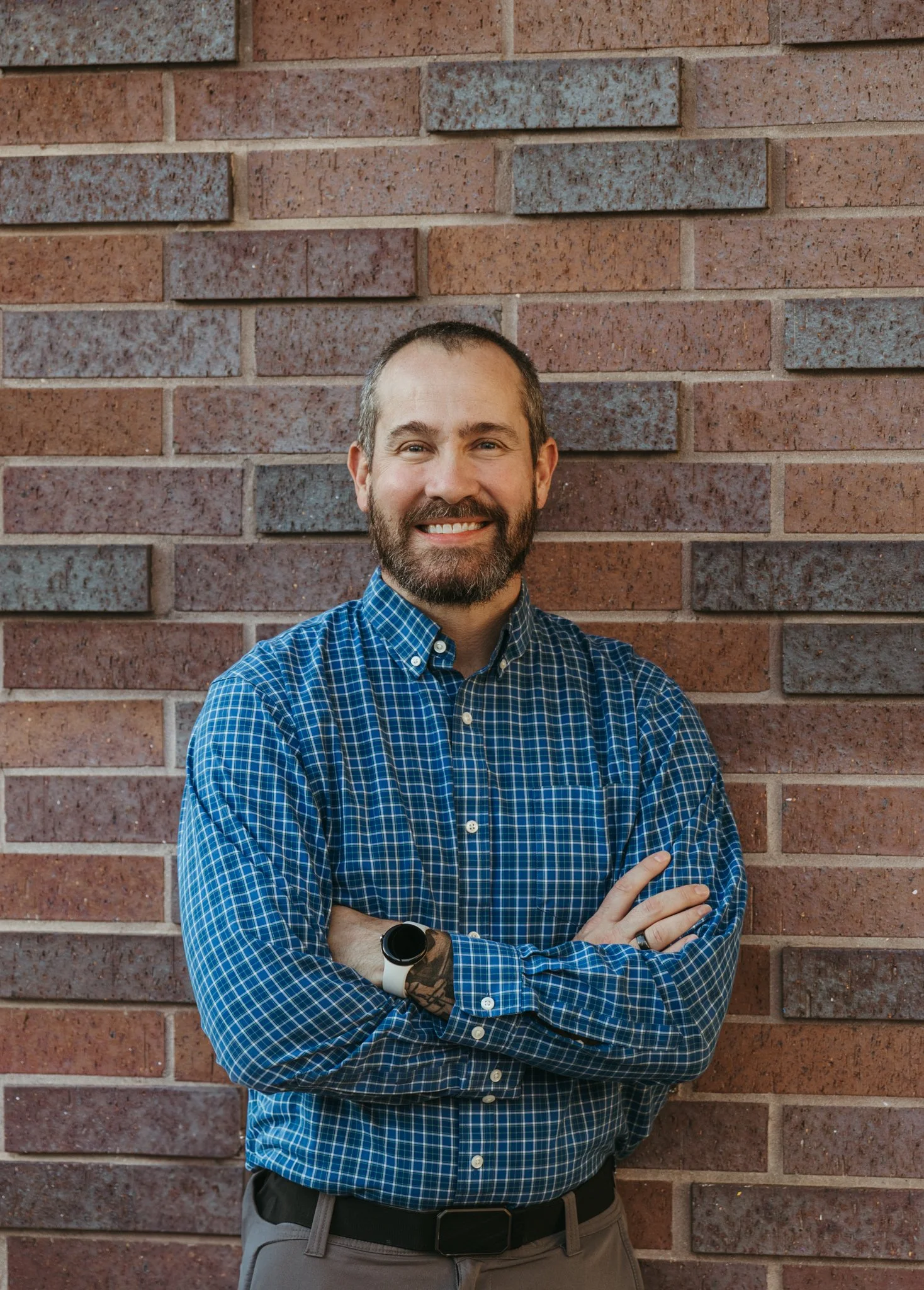 A man with short hair and a beard smiling with his arms crossed, standing in front of a brick wall, wearing a blue checkered shirt and a smartwatch.