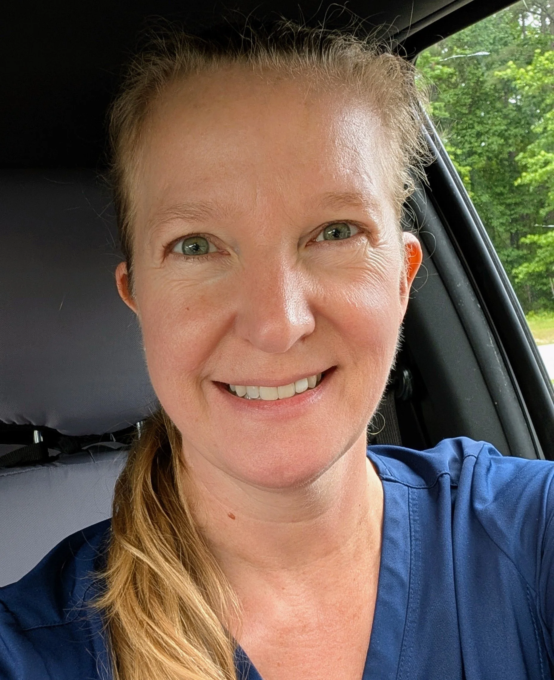 A woman smiling inside a vehicle, with trees visible through the window in the background.