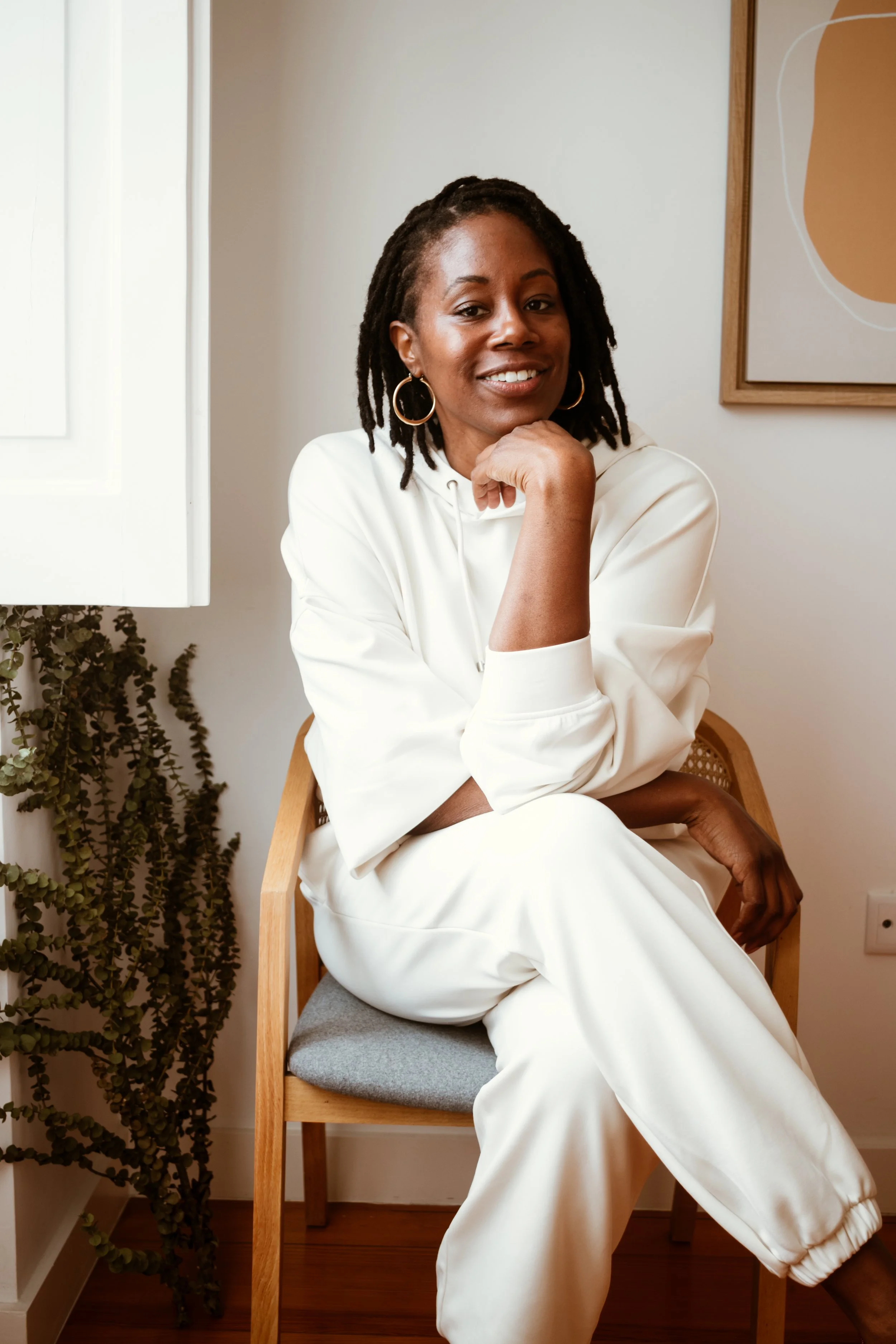 A woman with dreadlocks in a white hoodie and sweatpants sitting on a wooden chair in a cozy room with plants and abstract wall art.