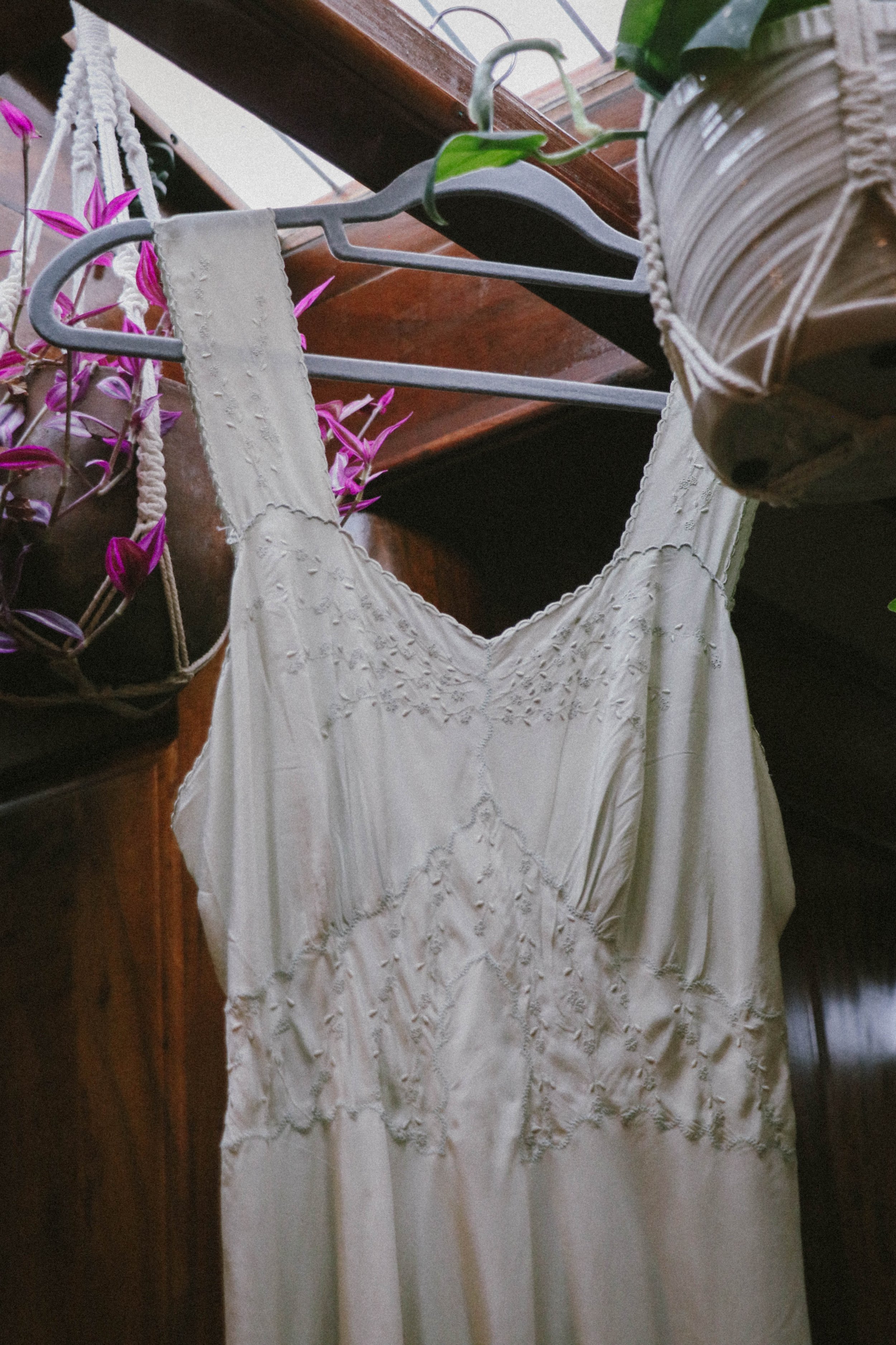 A white dress hanging on a black hanger on a wooden rack, with a purple plant in a pot nearby and a window above with natural light.
