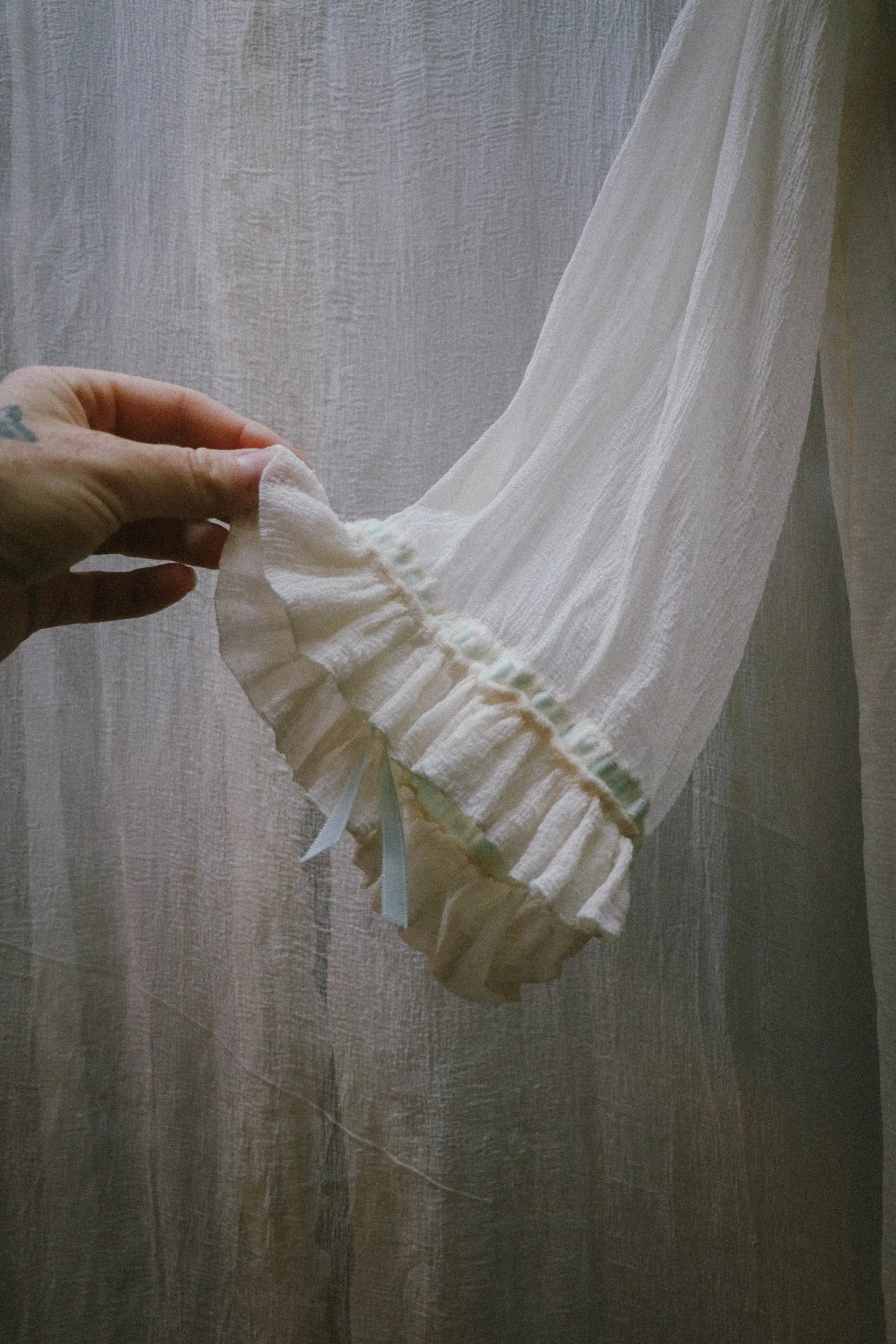 A person holding a white, long-sleeved fabric garment with ruffled cuffs in front of a textured, light-colored curtain.