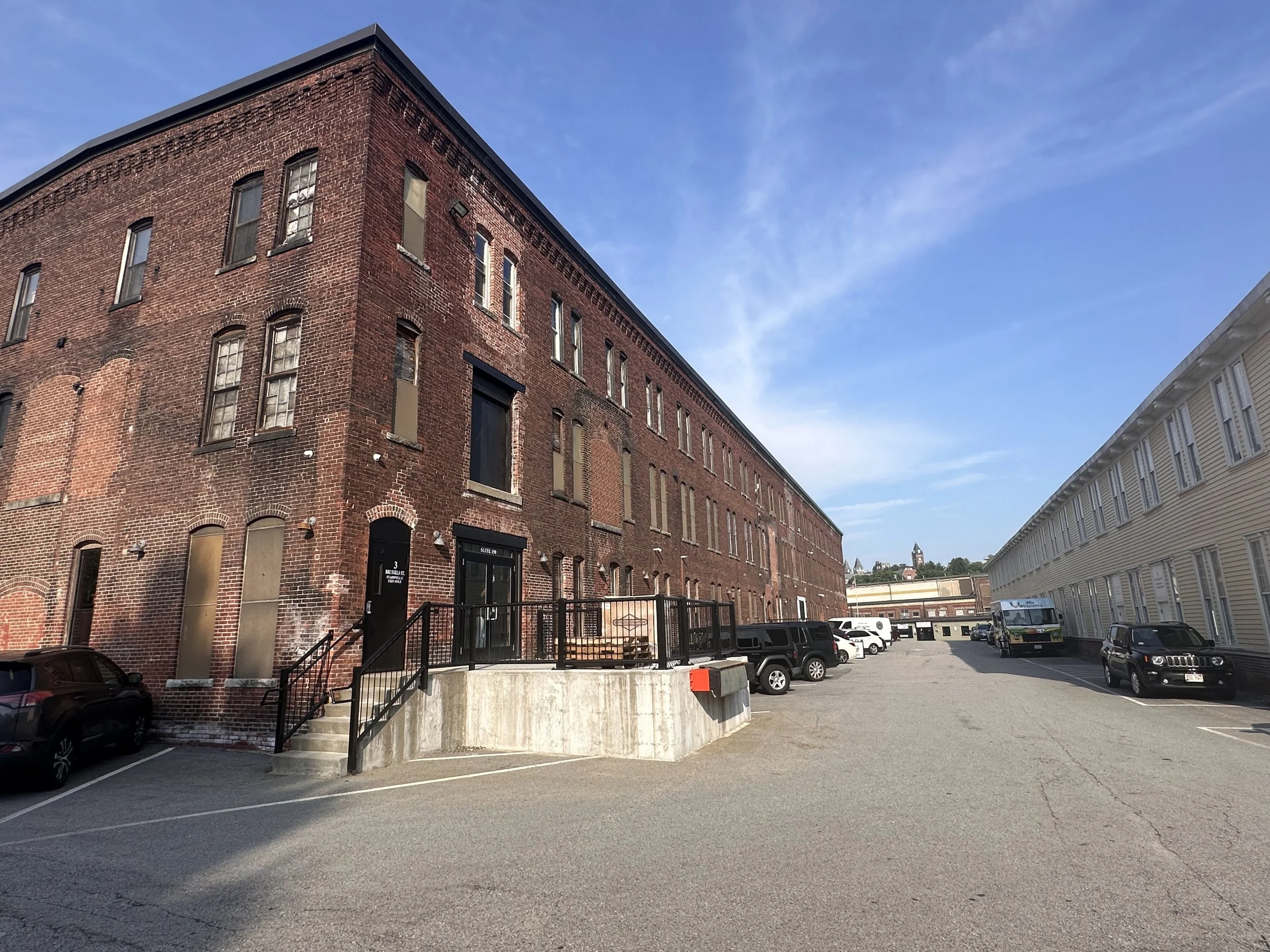 A parking lot adjacent to a large red brick building on the left and a white building on the right under a blue sky.