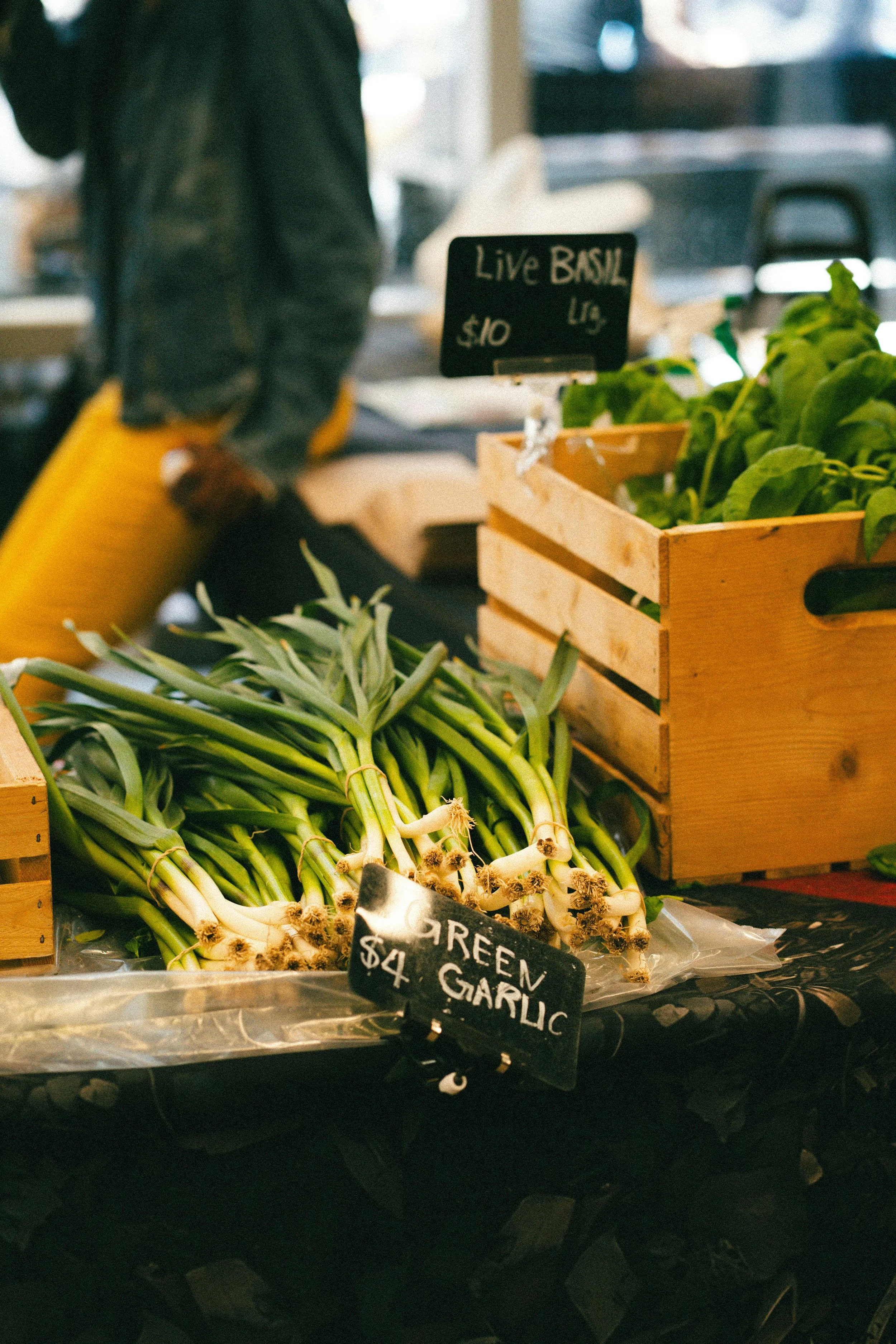 Fresh green garlic bunches on a market stall with a small black sign reading 'GREEN GARLIC $4' in front of them, and a crate of leafy greens nearby.