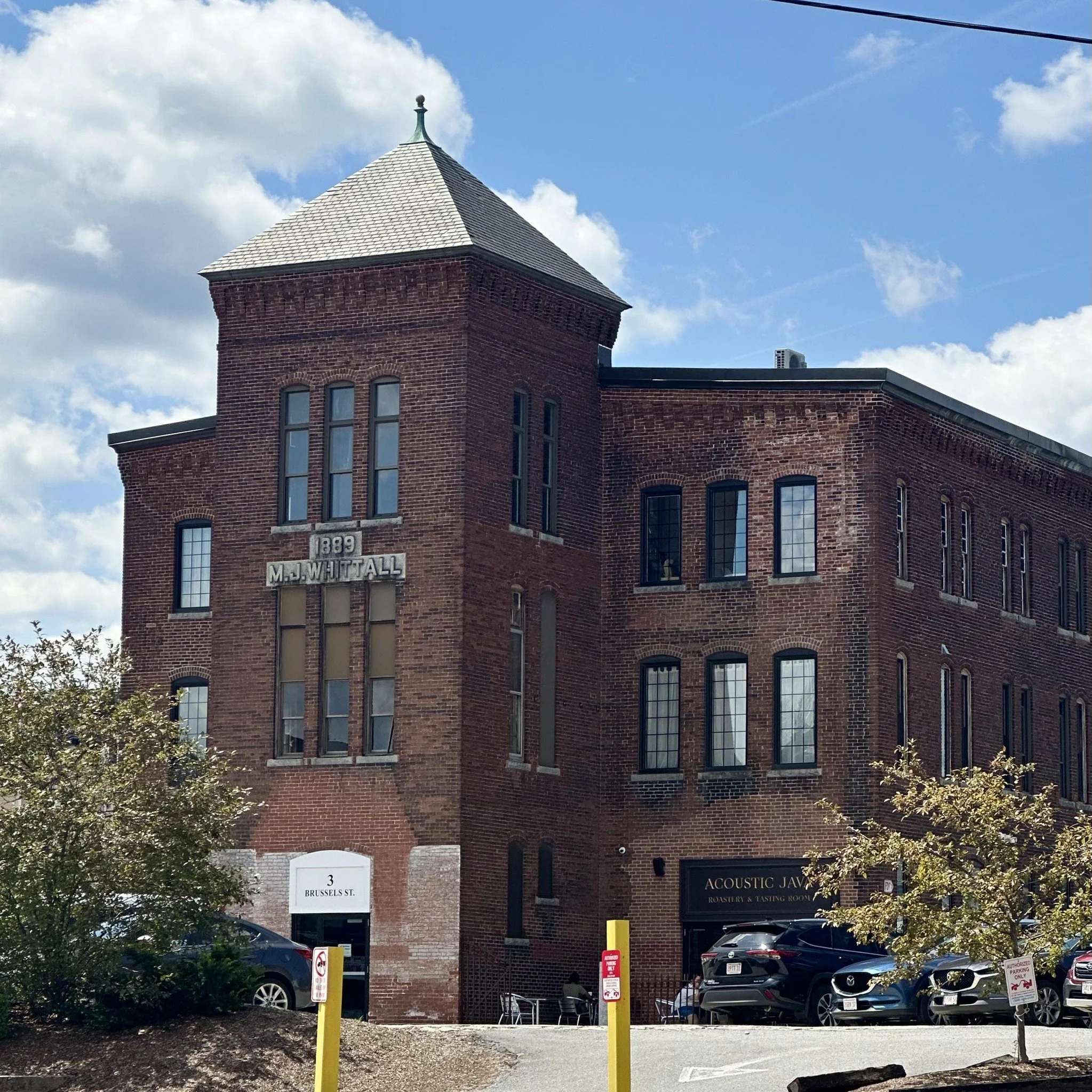 A three-story brick building with a tower on the left side, dated 1889, with tall narrow windows and a sign for Acoustic Java on the ground floor. The building is beside a parking lot with cars and trees, bright blue sky with white clouds in the background.