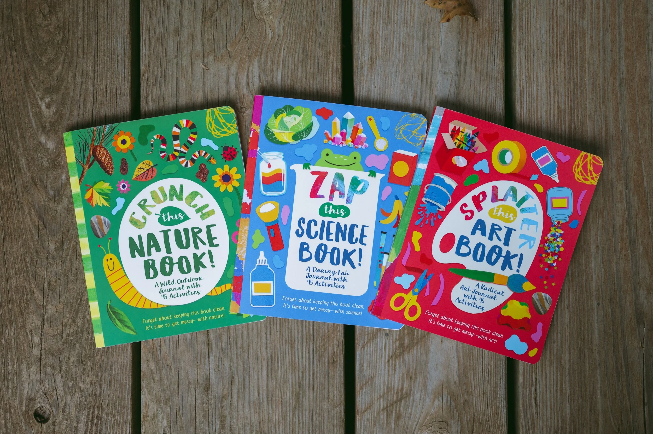 Three colorful activity books laid out on a wooden surface. The books are titled "Crunch this Nature Book!," "Zap this Science Book!," and "Splash this Art Book!" with vibrant illustrations on their covers.