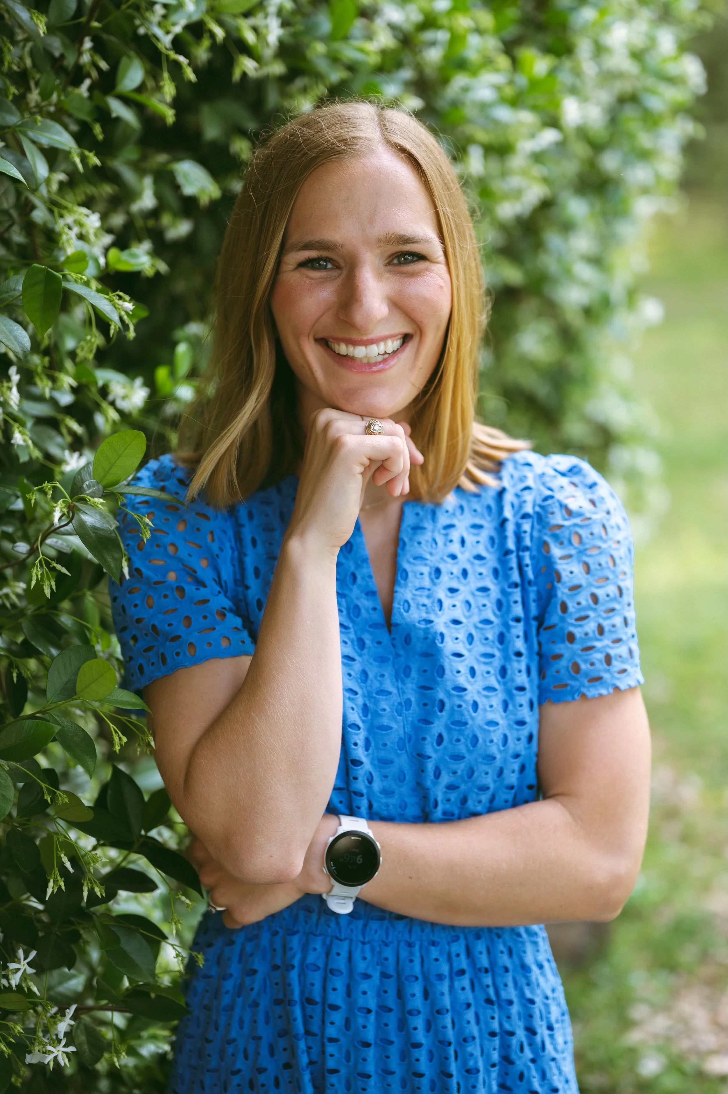 A woman with shoulder-length blonde hair smiling and posing outdoors, wearing a blue eyelet dress, a watch on her left wrist, and a ring on her right hand, standing next to green foliage.