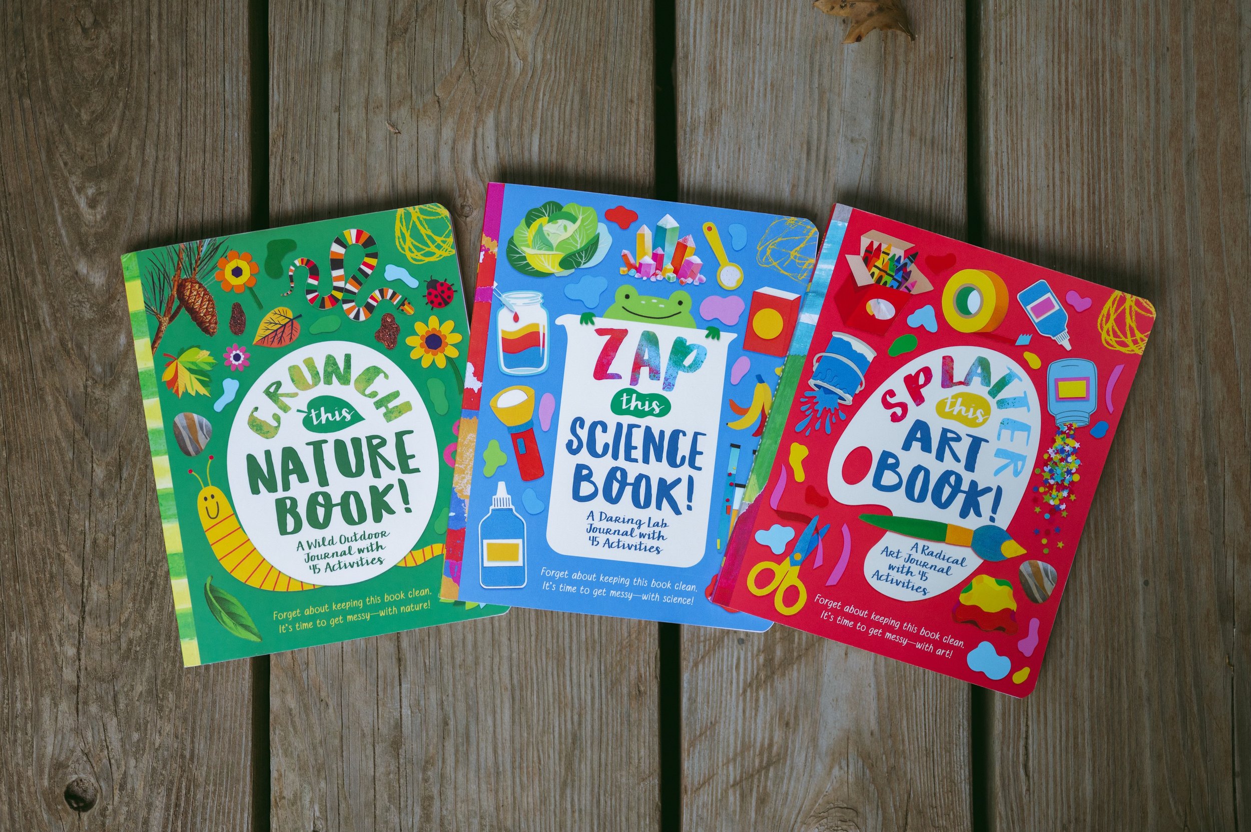 Three colorful children's activity books on a wooden surface: one titled "Crunch this Nature Book!", another "Zap this Science Book!", and the third "Splash this Art Book!"