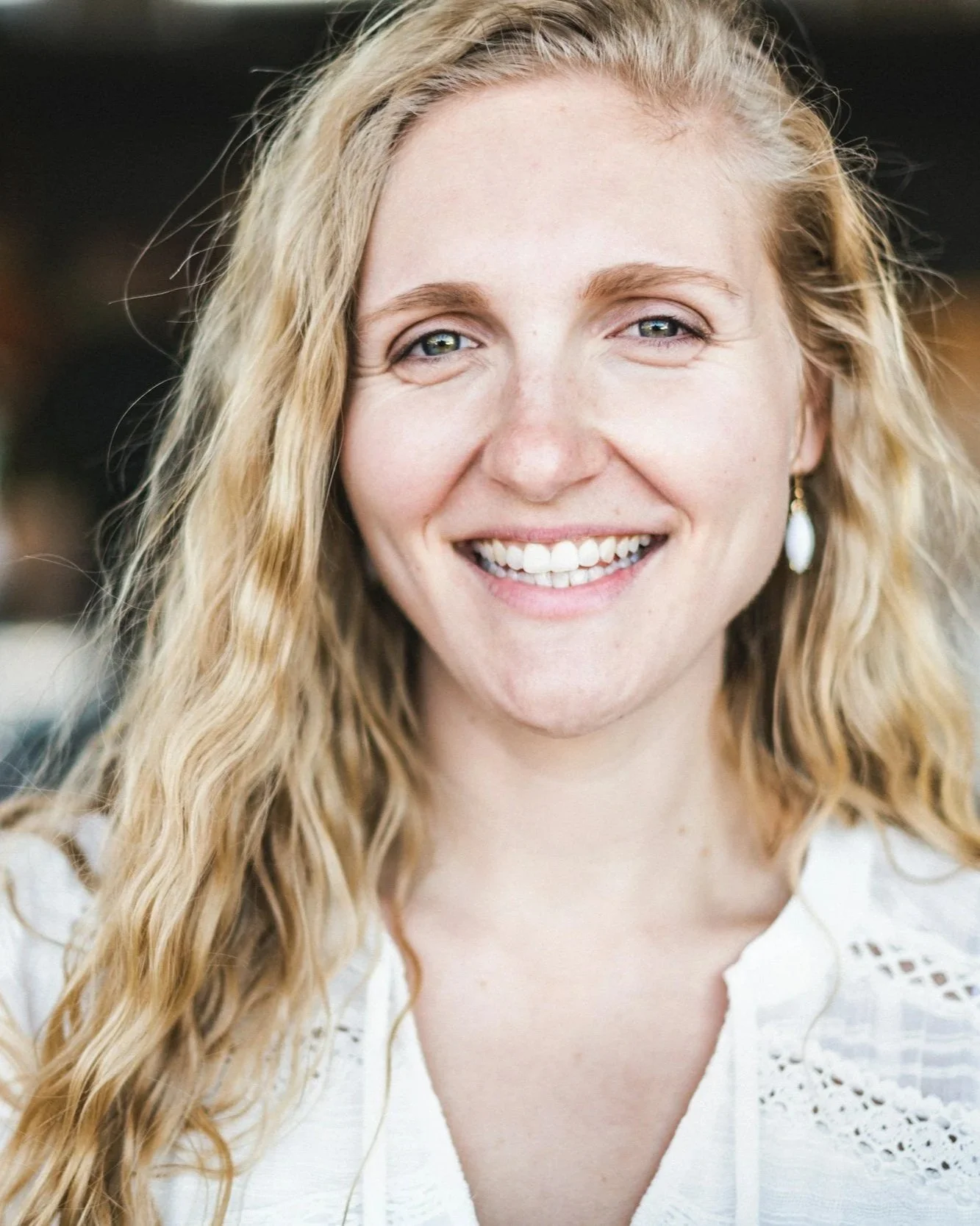 Nahaufnahme einer lachenden, blonde Frau mit Locken, weißen Zähnen und blauen Augen, trägt eine weiße Bluse mit Lochmuster und Ohrringe.