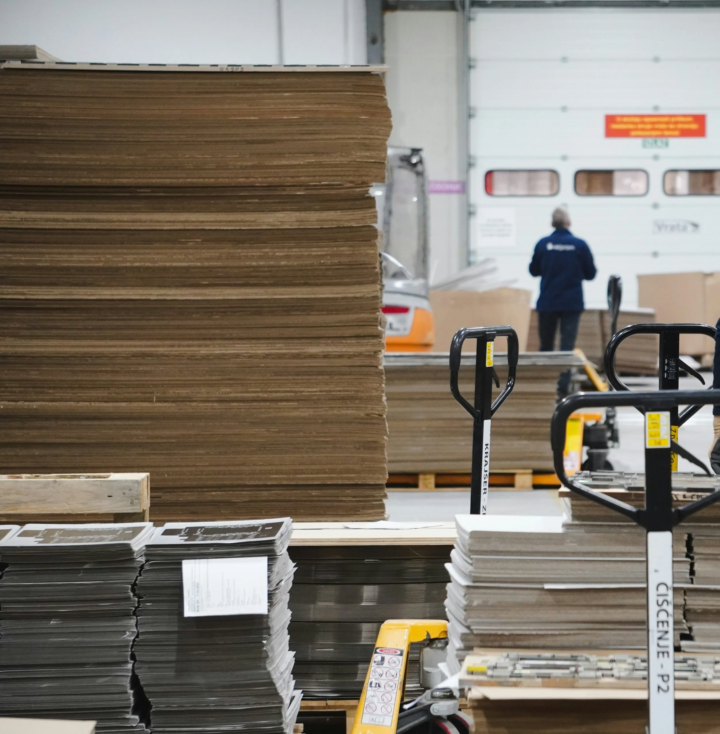 Stacks of cardboard and paper in a warehouse with a person in the background.
