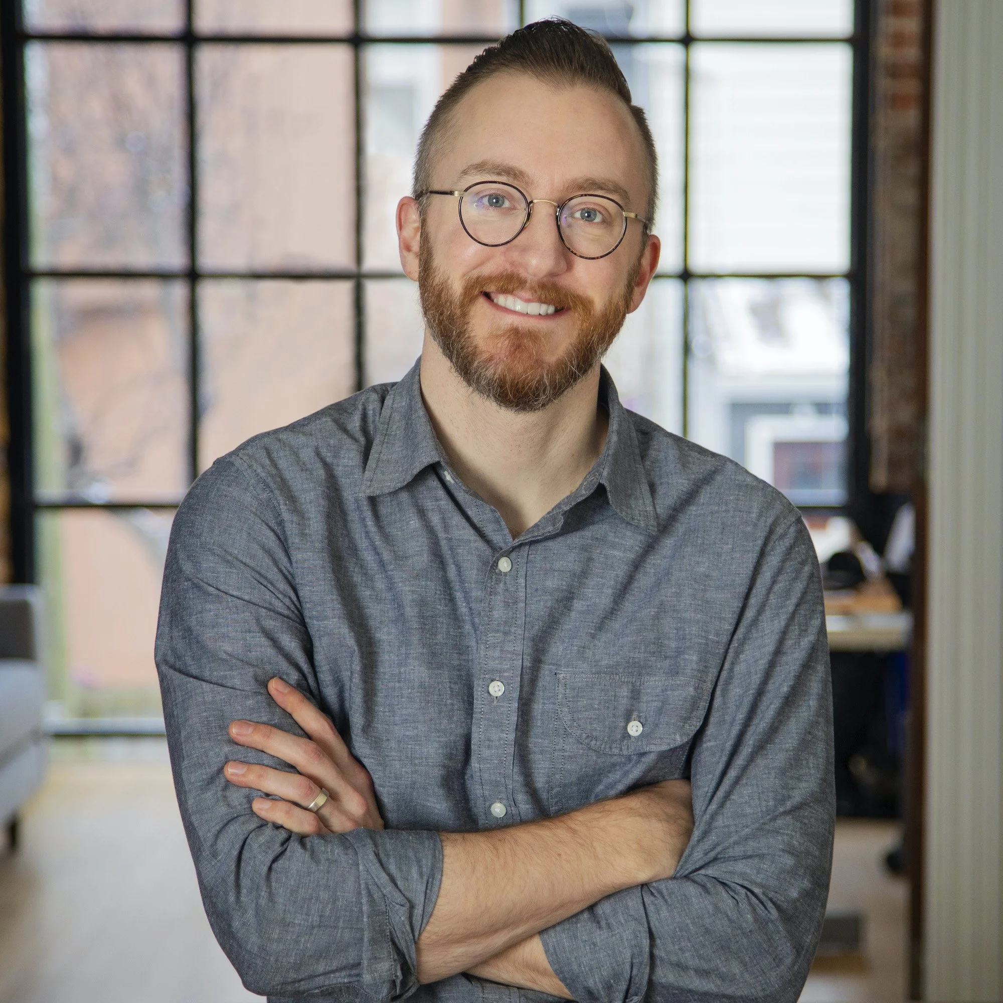A smiling man with glasses and a beard, wearing a gray button-up shirt, standing with arms crossed in a modern office with large windows.