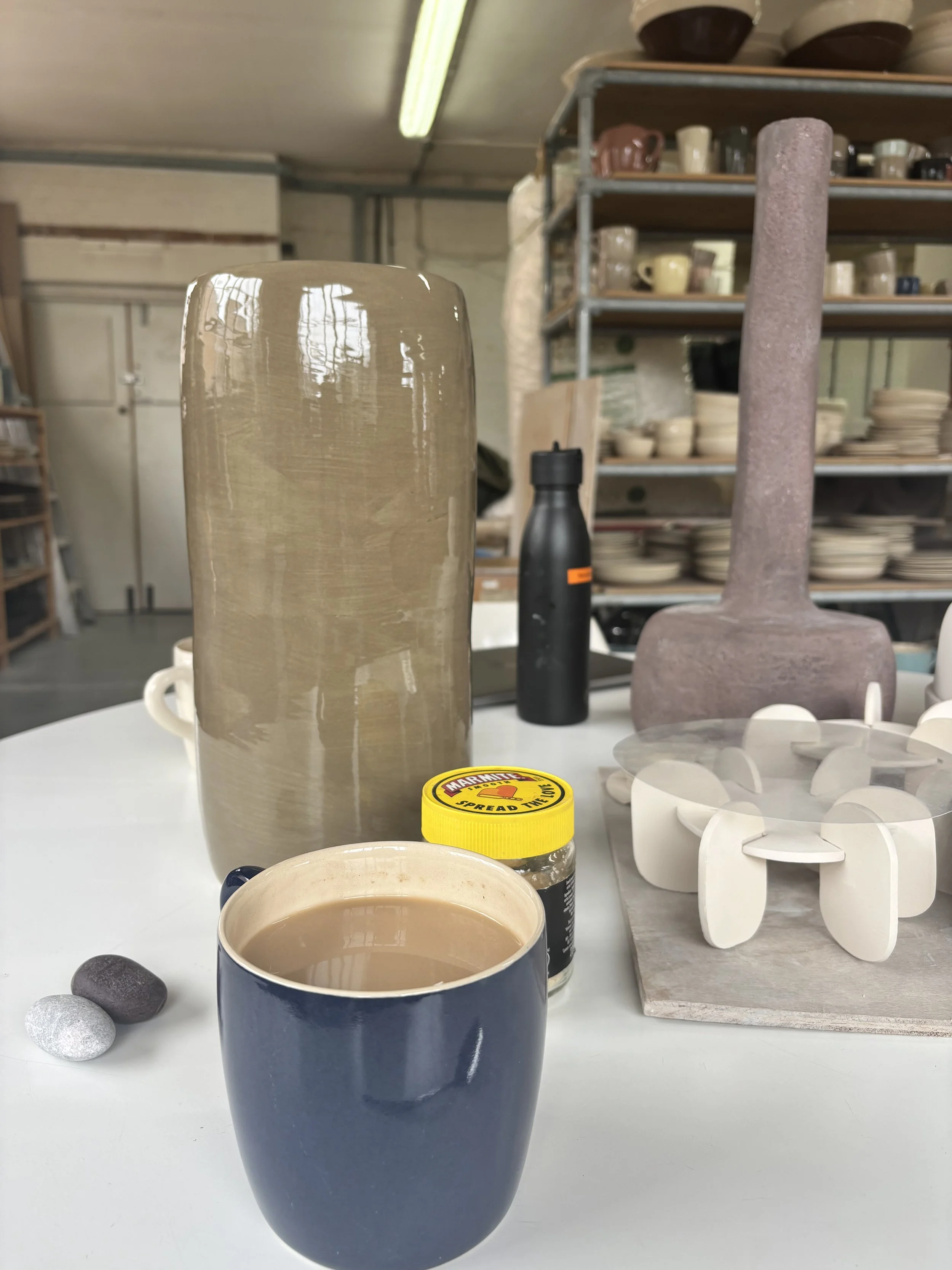 A blue mug filled with coffee, a small jar of Marmite, decorative stones, a ceramic vase, and a wooden tray with white ceramic flower-shaped objects on a white table. In the background, shelves with various cups and bowls in a pottery studio.