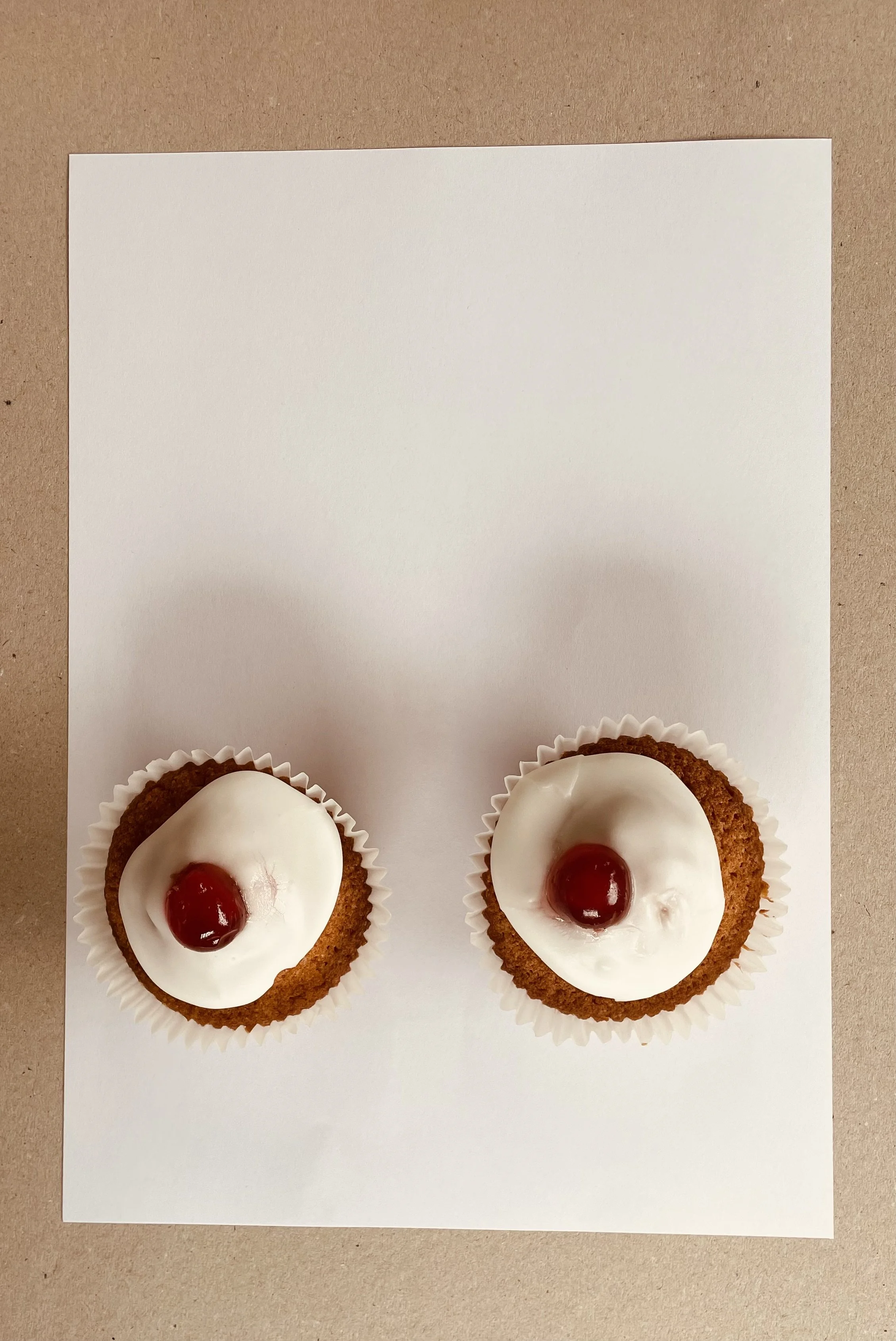 Two cupcakes with white frosting and a red cherry on top, placed on a white sheet of paper on a brown surface.