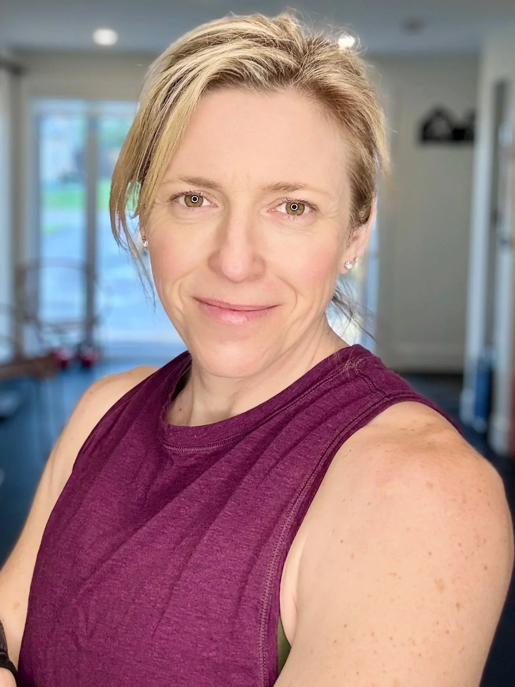 A woman with short blonde hair and brown eyes smiling, wearing a sleeveless maroon top, inside a room with large windows in the background.