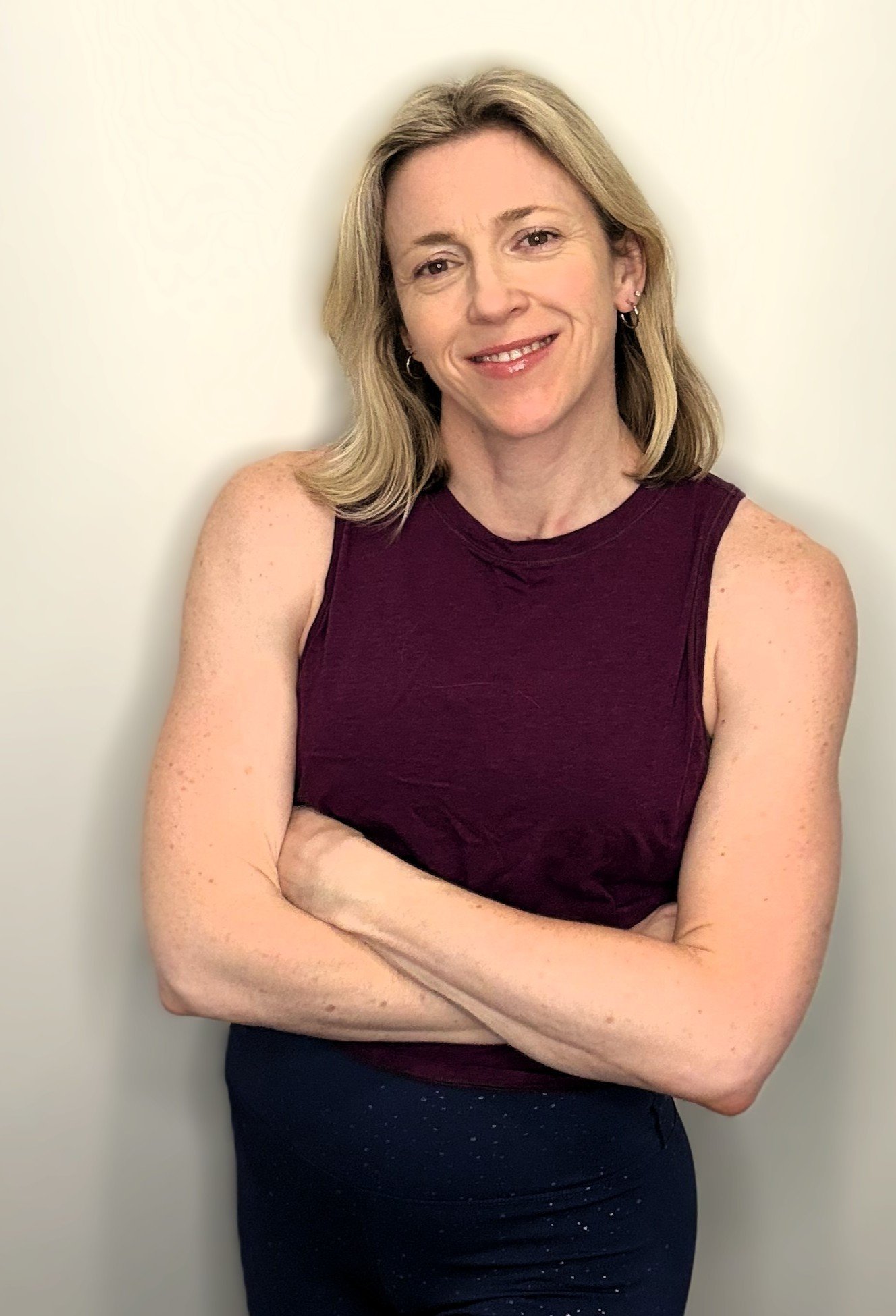 A woman with shoulder-length blonde hair smiling, wearing a sleeveless maroon top and dark pants, crossing her arms, standing against a plain light-colored background.