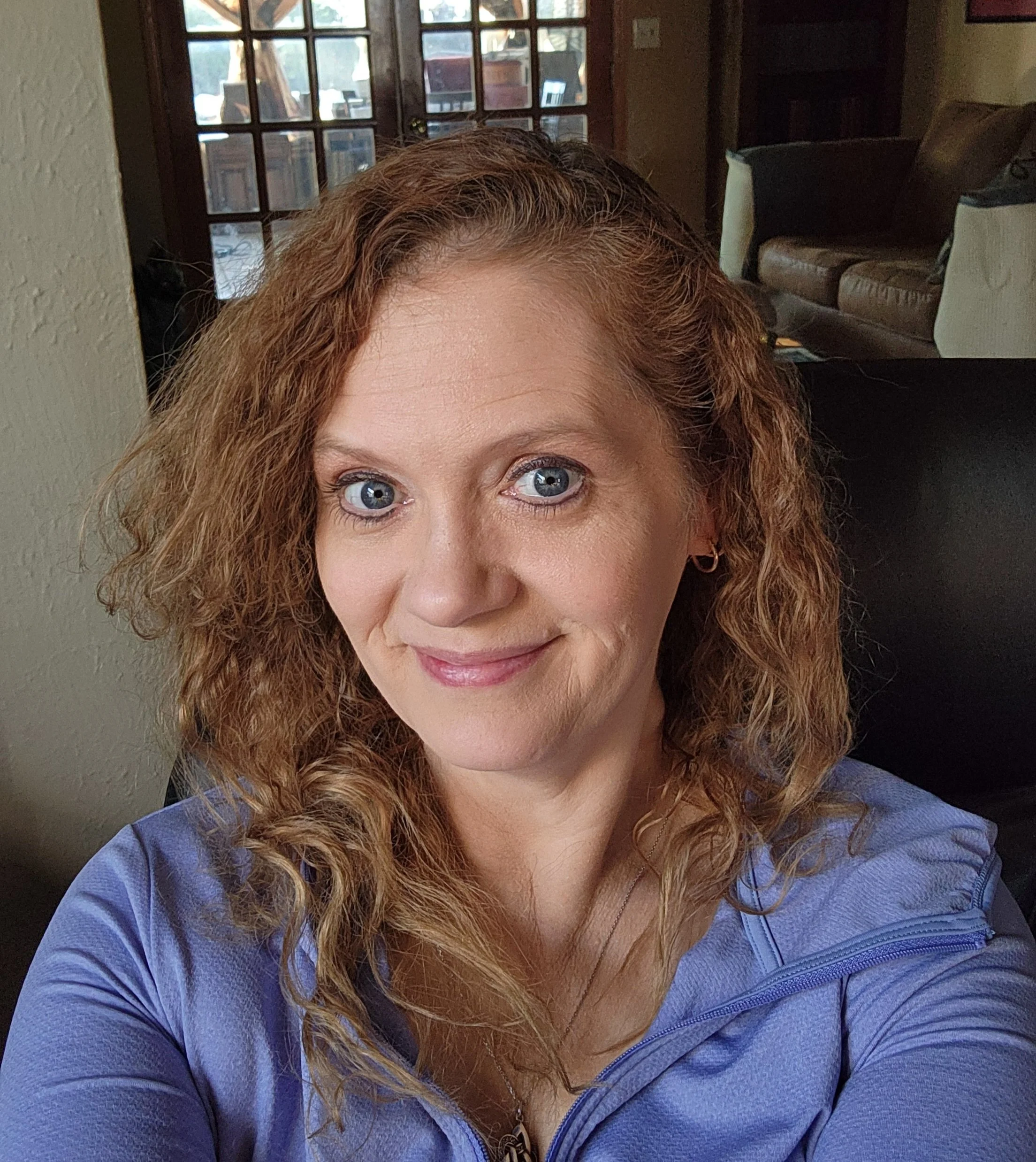 Close-up of a woman with curly red hair and blue eyes, sitting indoors with a window in the background. She is smiling slightly and wearing a blue top.