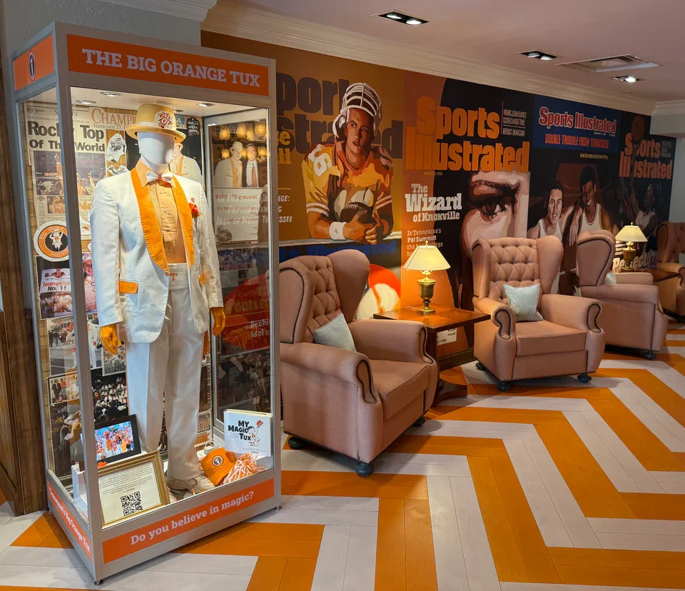 Display case with a mannequin dressed in a white tuxedo with orange accents, surrounded by sports memorabilia and magazines. The room has pink armchairs, side tables with lamps, and walls decorated with colorful sports magazine covers.