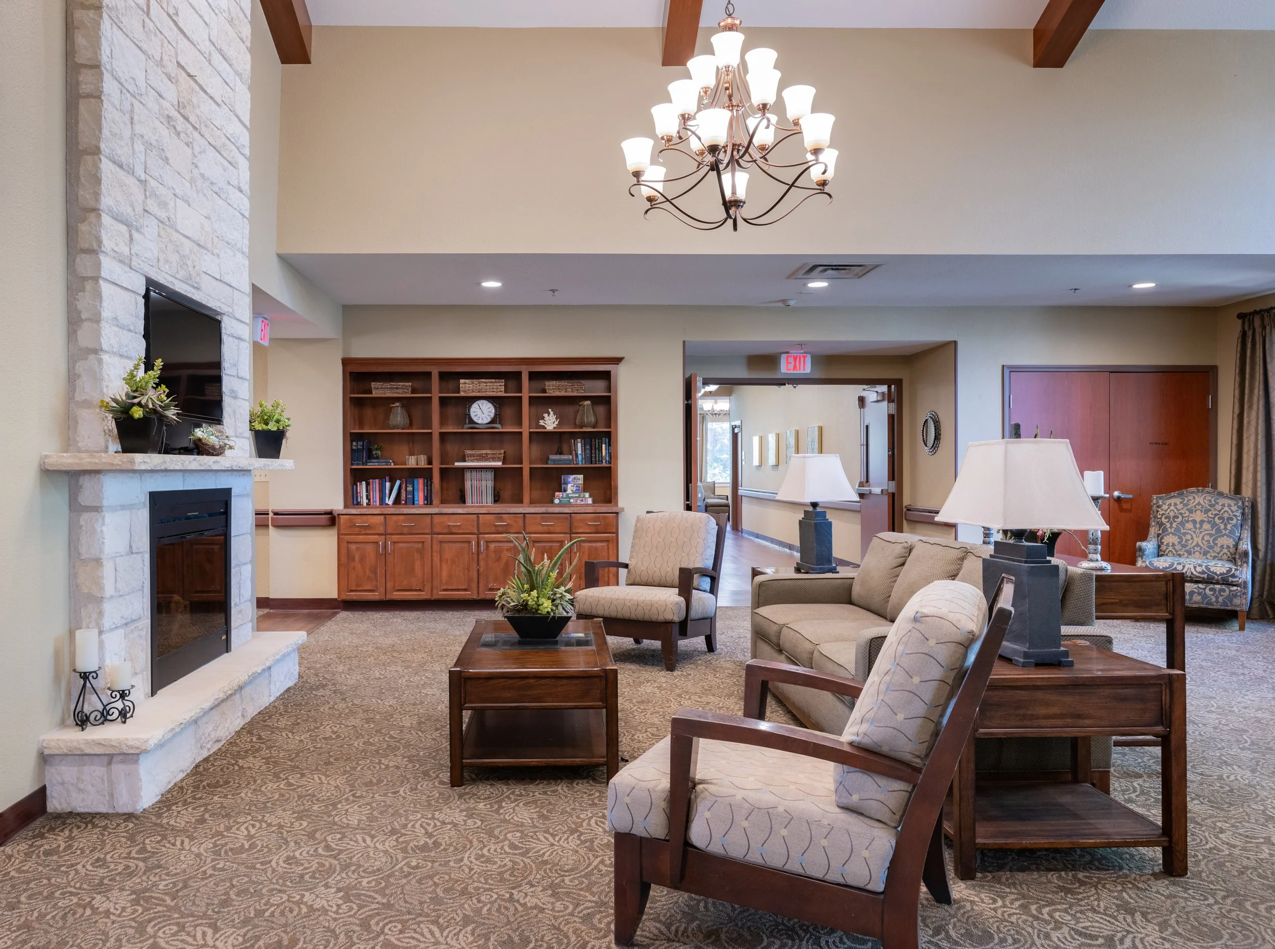 A cozy nursing facility lobby with a stone fireplace, wooden bookshelf, beige seating, coffee table with a plant, and a chandelier hanging from the ceiling.