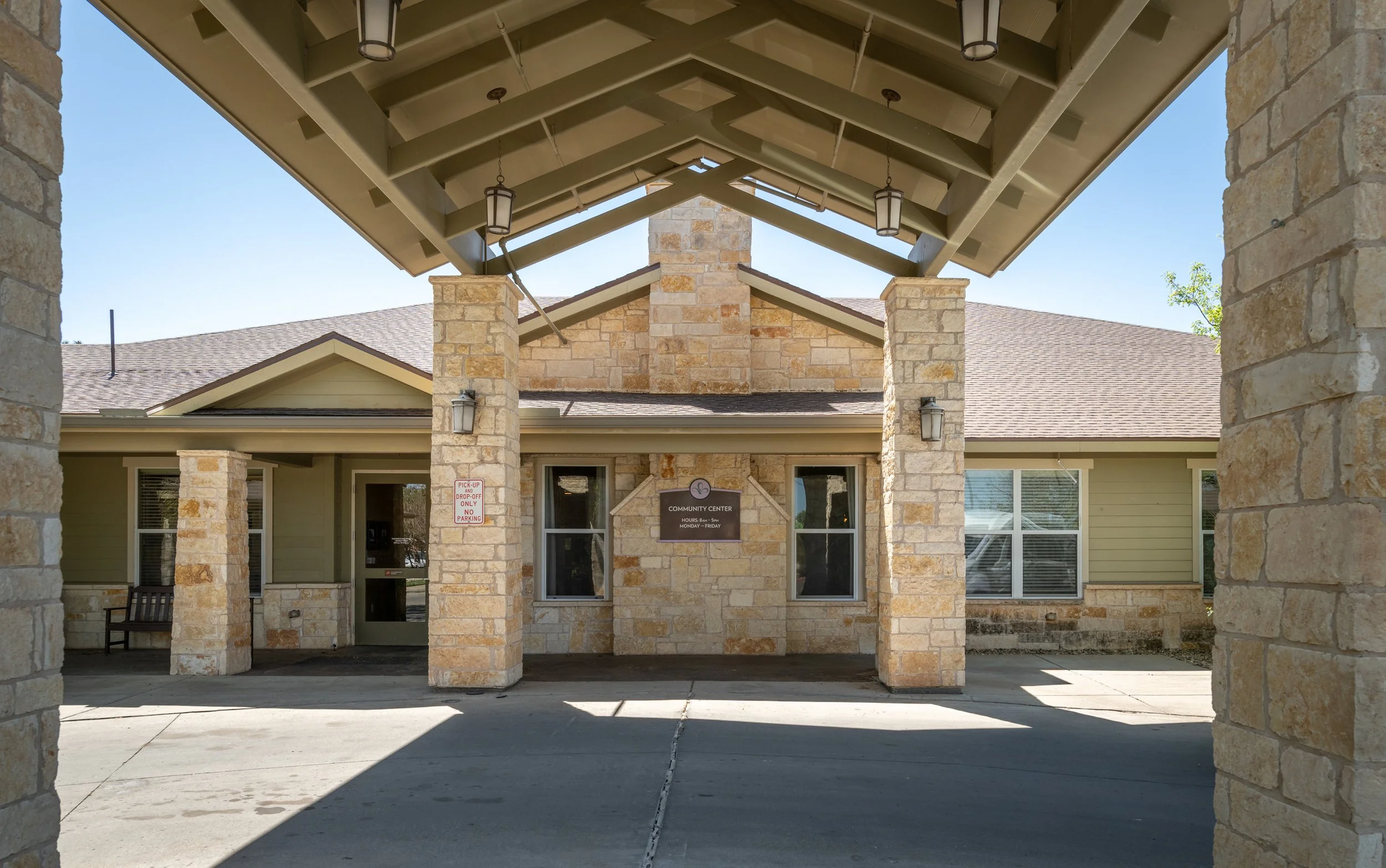 View of a nursing home entrance with stone columns and a covered driveway, sign indicating hours on a sunny day.