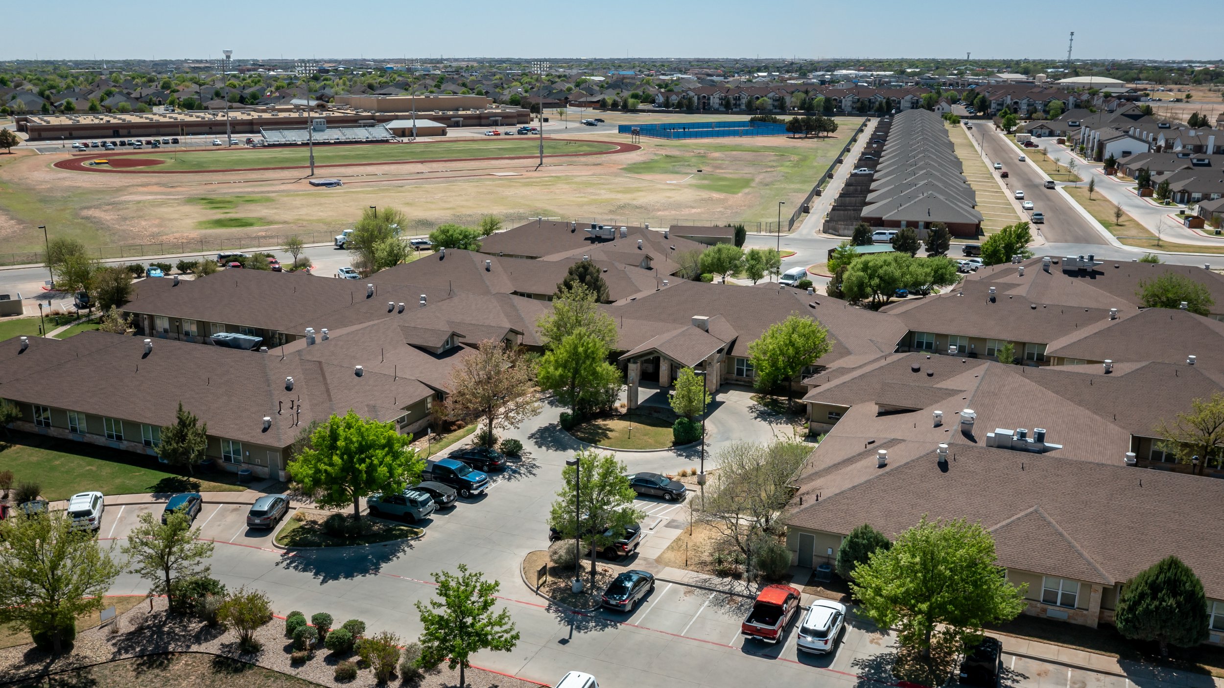 Aerial view of skilled nursing facility with brown roofs, surrounded by trees and parking lots, with a sports field and housing in the background.