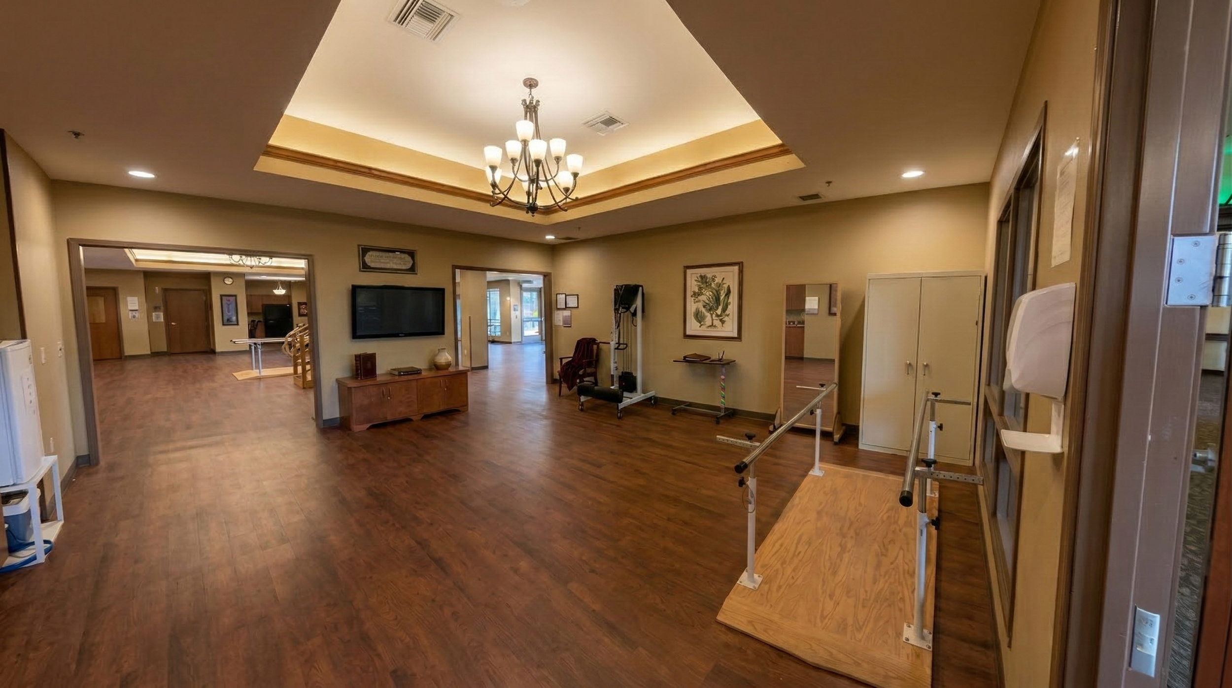 Indoor lounge area with wooden floors, beige walls, a chandelier, wall-mounted TV, artwork, indoor exercise equipment, and a doorway leading to an outdoor patio.