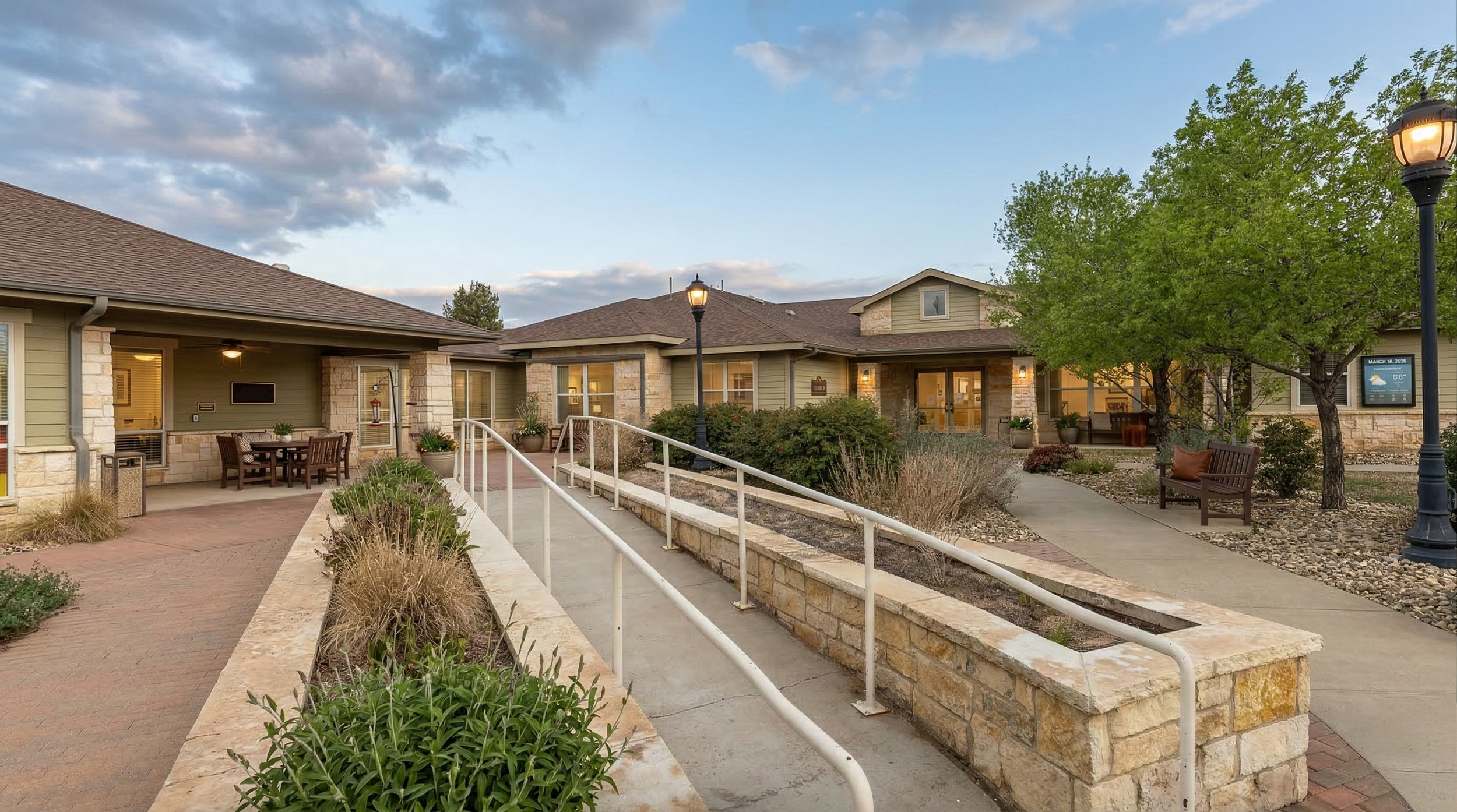 Exterior view of a skilled nursing facility at sunset with trees, bench, lamp posts, landscaped gardens, and accessible ramp