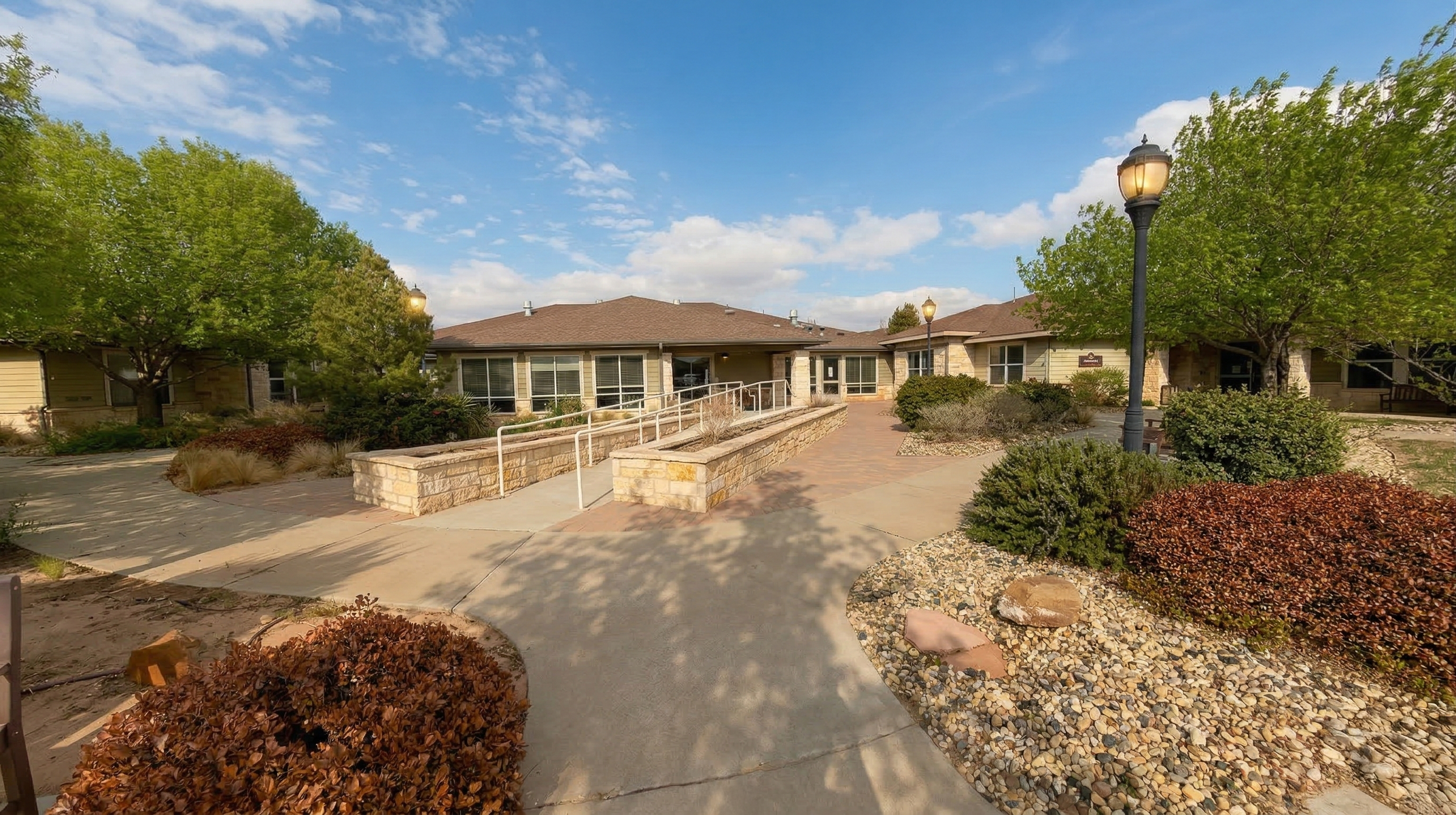 A modern post-acute care facility with large windows, surrounded by landscaped gardens, paved walkways, trees, street lamps, and clear blue sky.