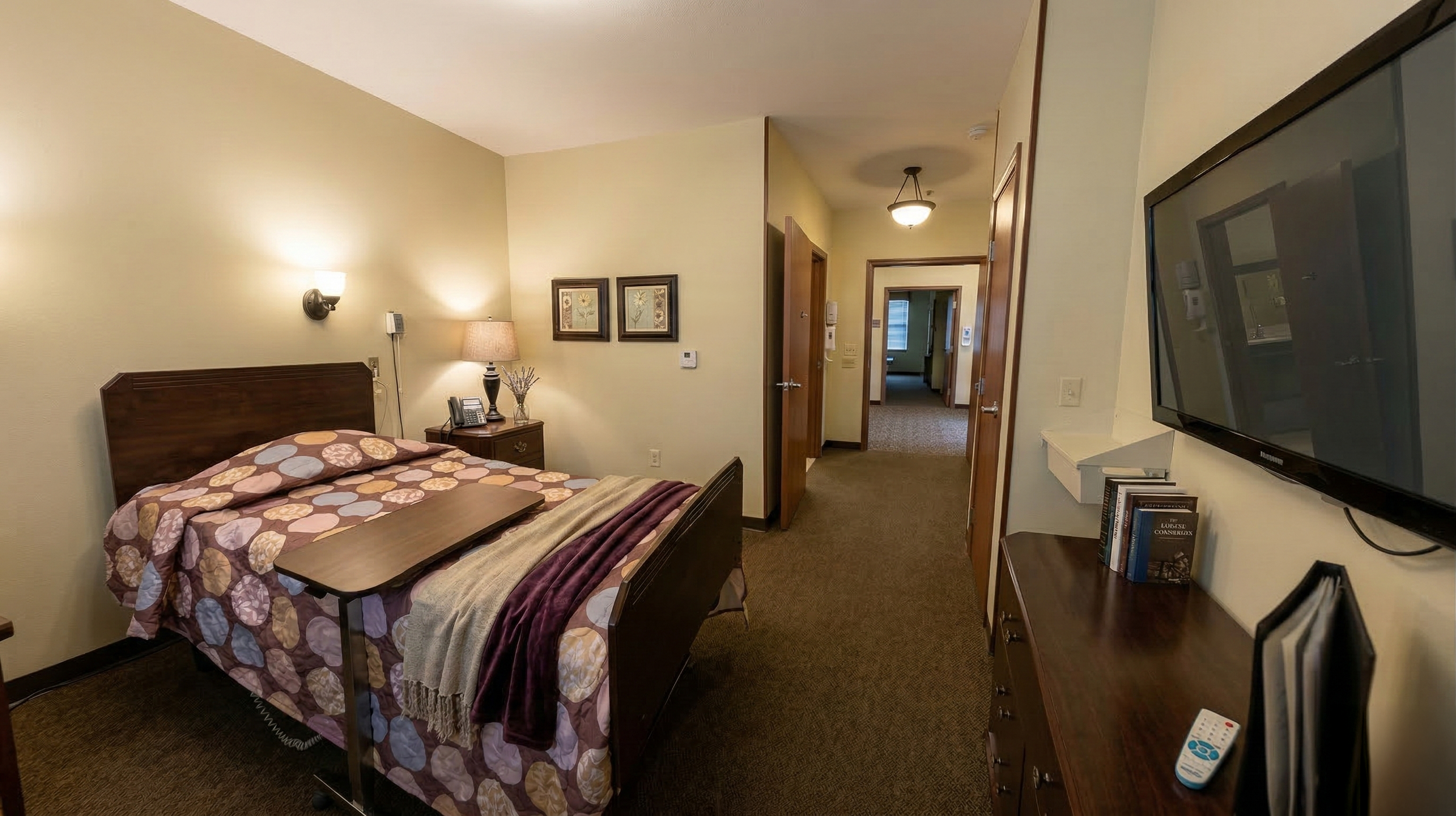Luxury private hospice suite with bed, nightstand with a lamp and phone, TV mounted on the wall, and a dresser with books, in a hallway with open doors leading to other rooms.