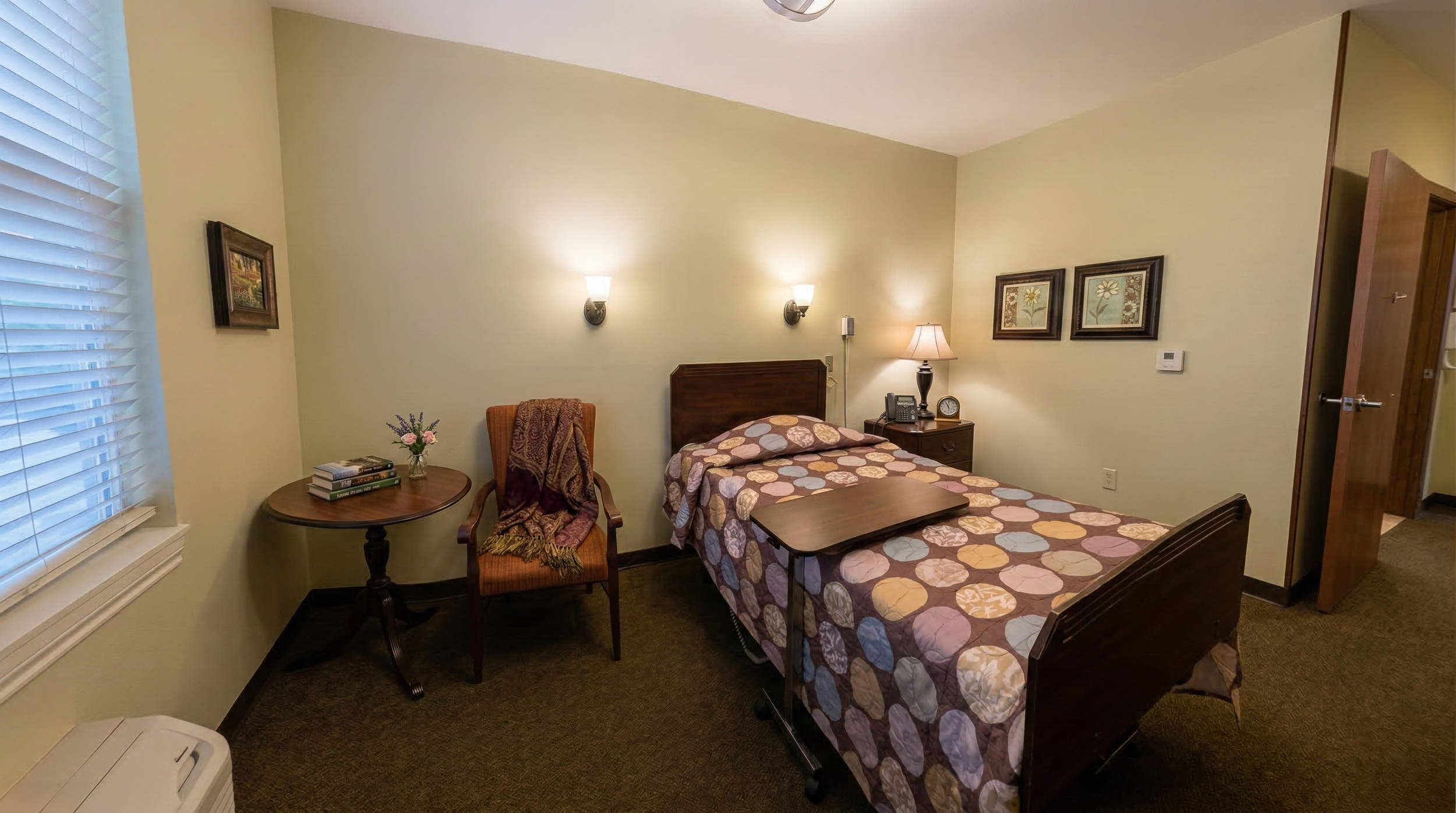Spacious resident room with a bed, bedside table with lamp and clock, office chair with blanket, small round table with flowers and books, framed art on the walls, window with blinds, and carpeted floor.