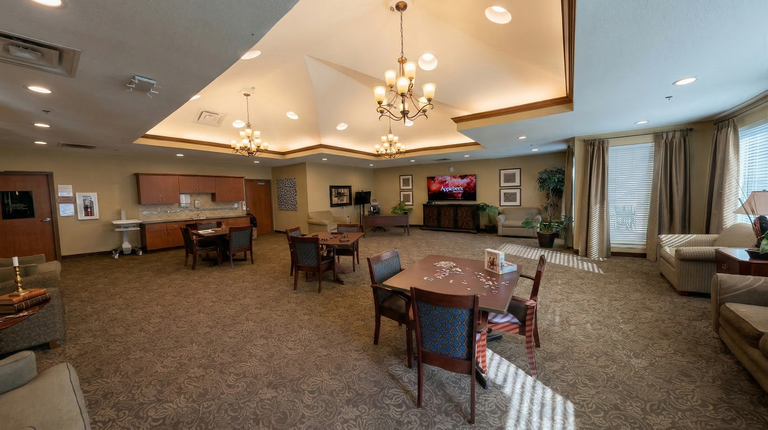 A spacious, well-lit common area with multiple seating areas, a large TV, and a kitchenette. The activity room features chandeliers, windows with curtains, and patterned carpeting.