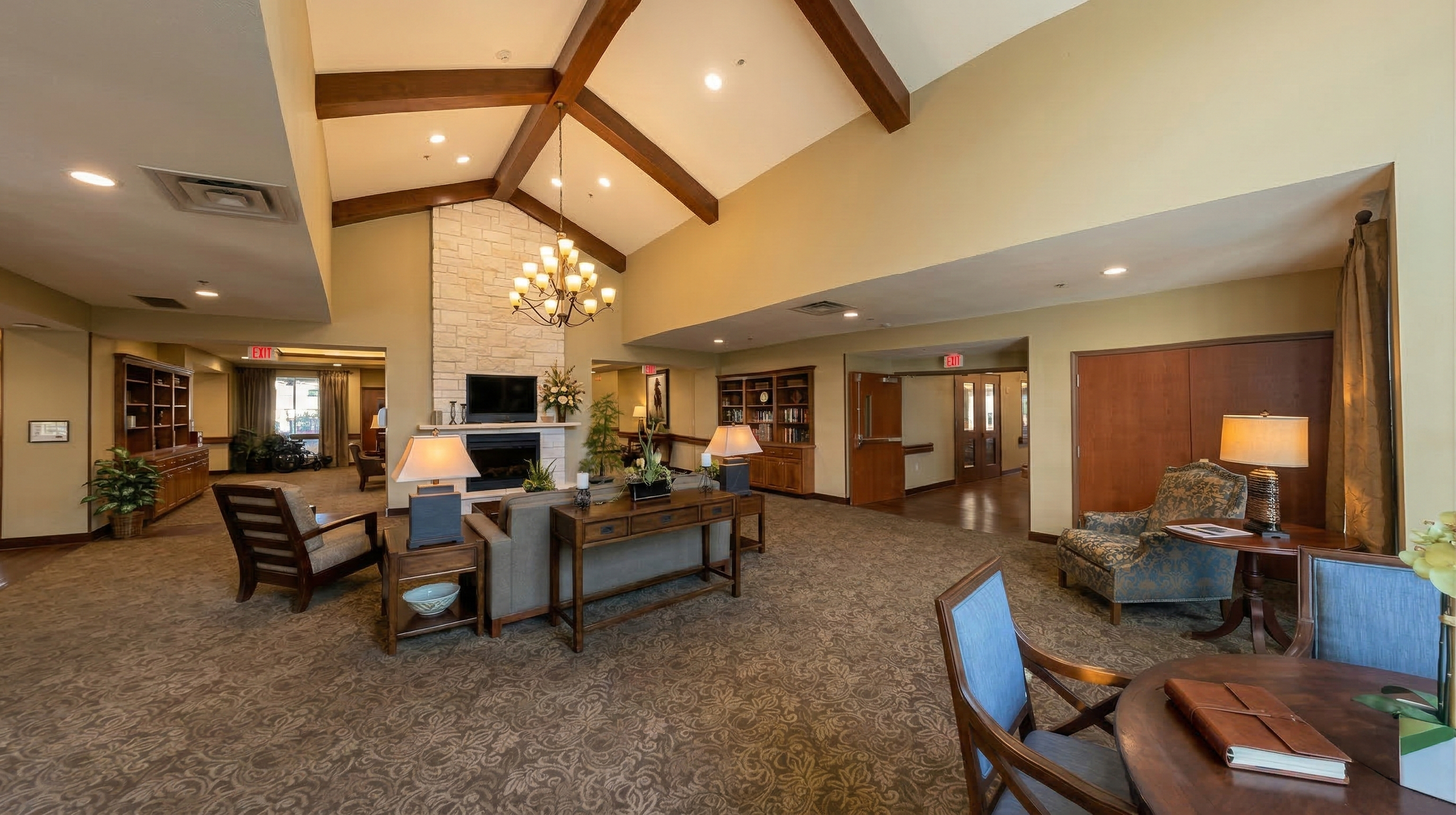 Spacious skilled nursing facility lobby with high ceilings, wooden beams, a stone fireplace with a television, multiple seating areas with lamps, and decorative plants.