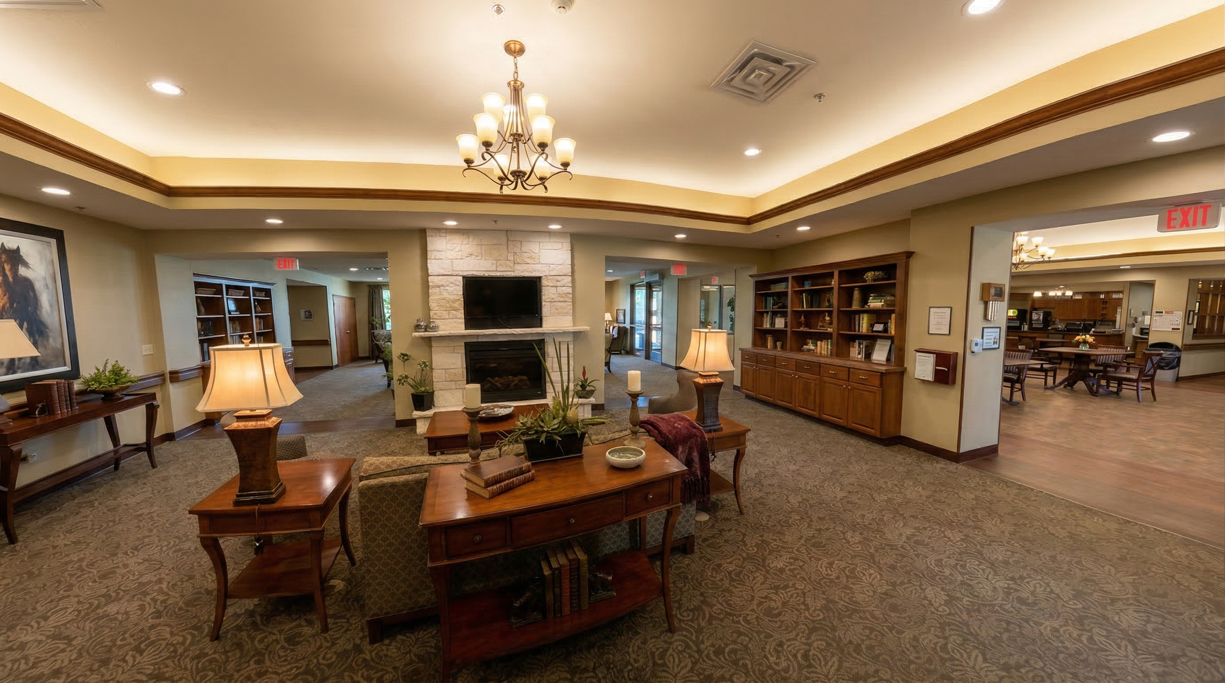 A spacious long term care facility lobby with a central seating area, a stone fireplace, a wall-mounted TV, wooden bookshelves, lamps, and a chandelier, with hallway entrances and an exit sign visible.
