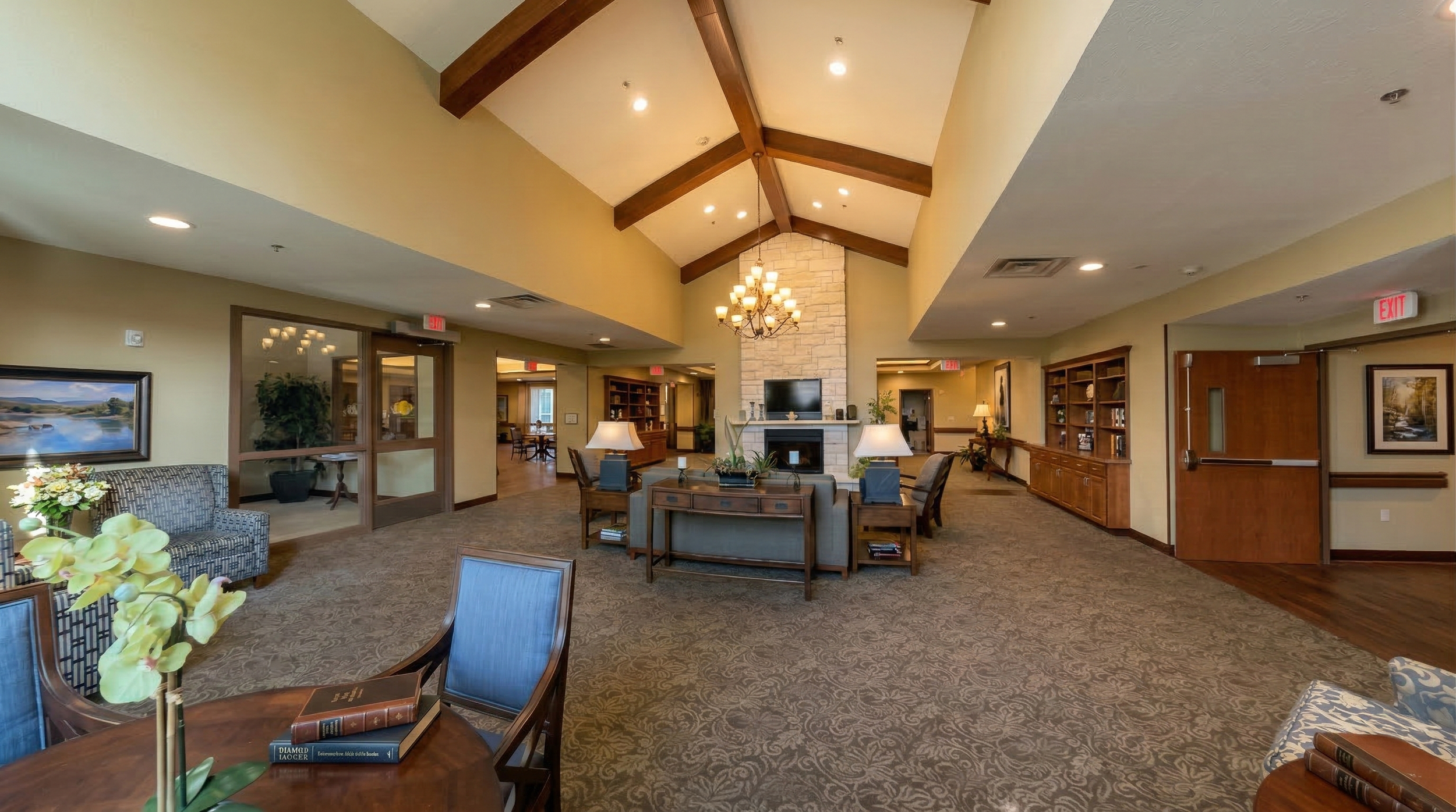 A spacious nursing home lobby with a high vaulted ceiling supported by wooden beams, a large chandelier, a fireplace, and comfortable seating areas. The room features warm colors, bookshelves, and framed artwork on the walls.