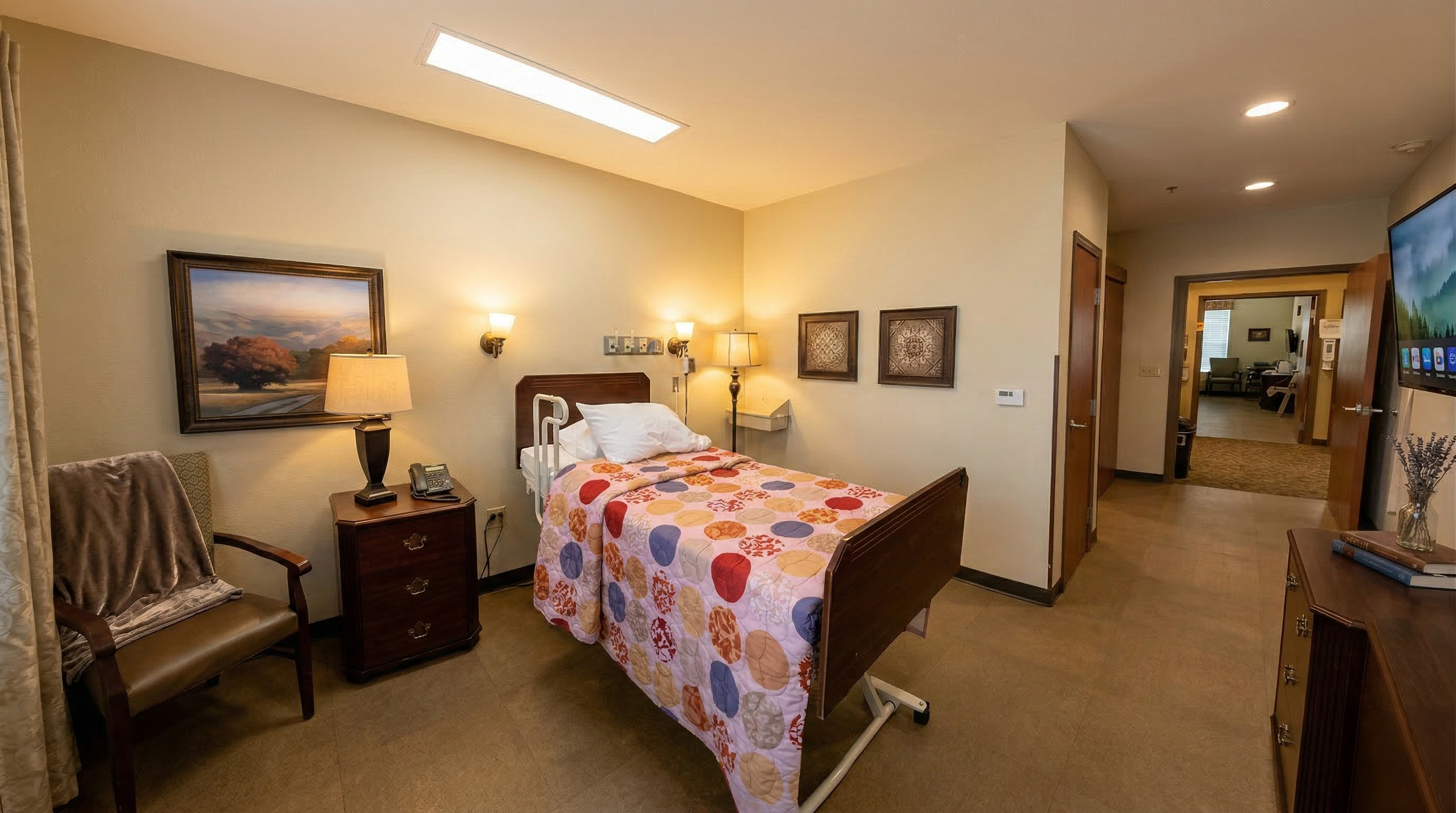 A care facility patient room with a bed, a chair, a side table, and wall-mounted lamps, with a view into another sitting area.