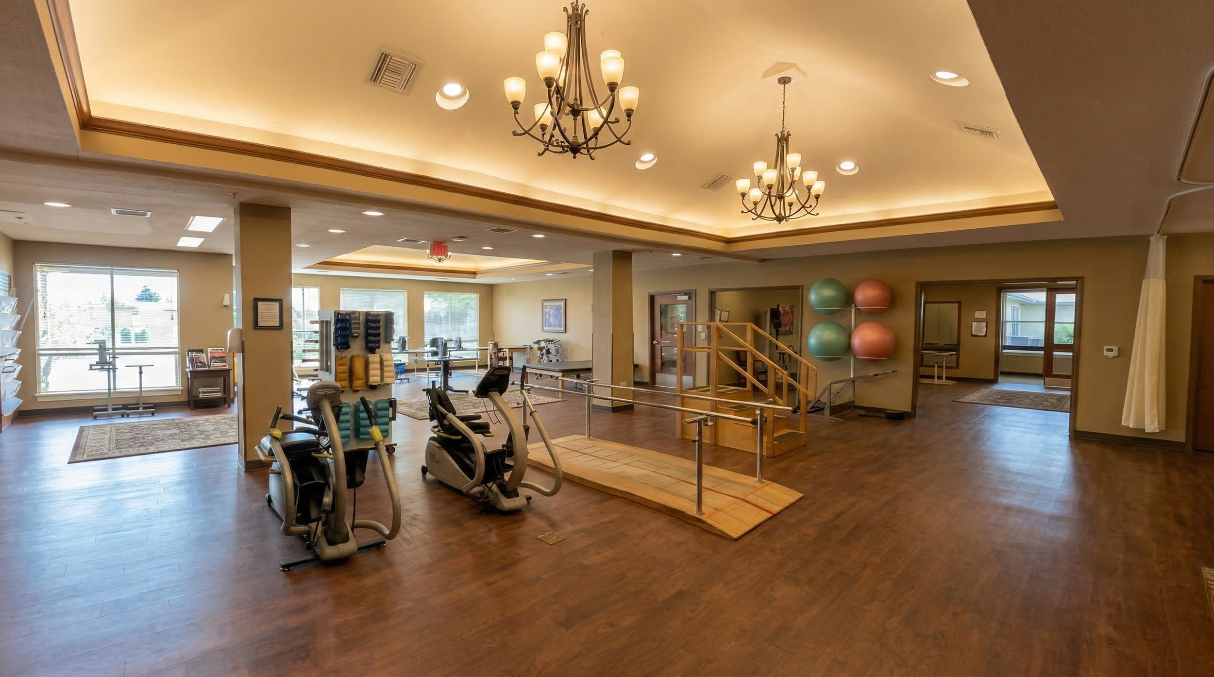 A spacious indoor rehab and therapy room with wooden floors, multiple gym equipment including stationary bikes, and large windows letting in natural light. There are chandeliers hanging from the ceiling and a wall with exercise balls.