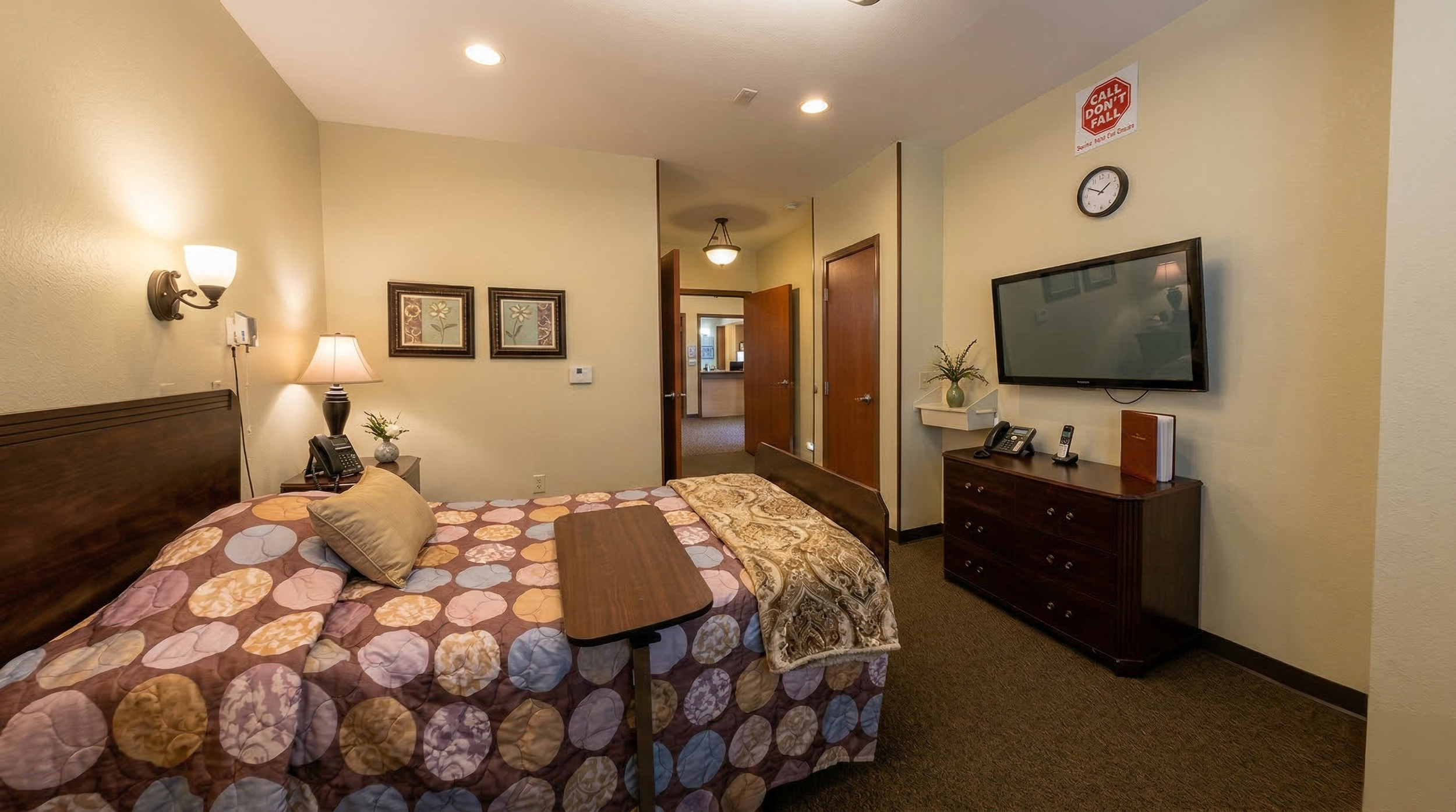 Senior care suite with a bed, a wooden nightstand with a lamp and phone, framed floral artwork on the wall, a dresser with phones and a TV, a wall clock, and a doorway leading to a hallway and other rooms.