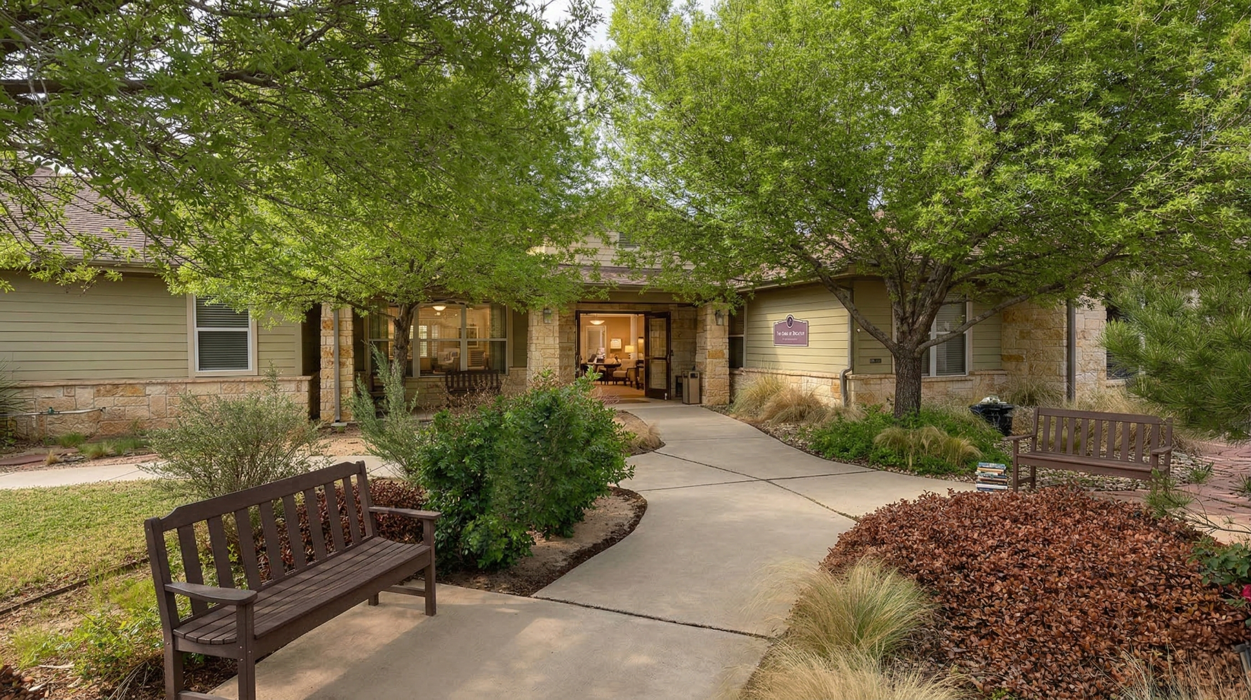 Exterior gardens of a nursing facility with a curved concrete walkway, benches, trees, and landscaped bushes.